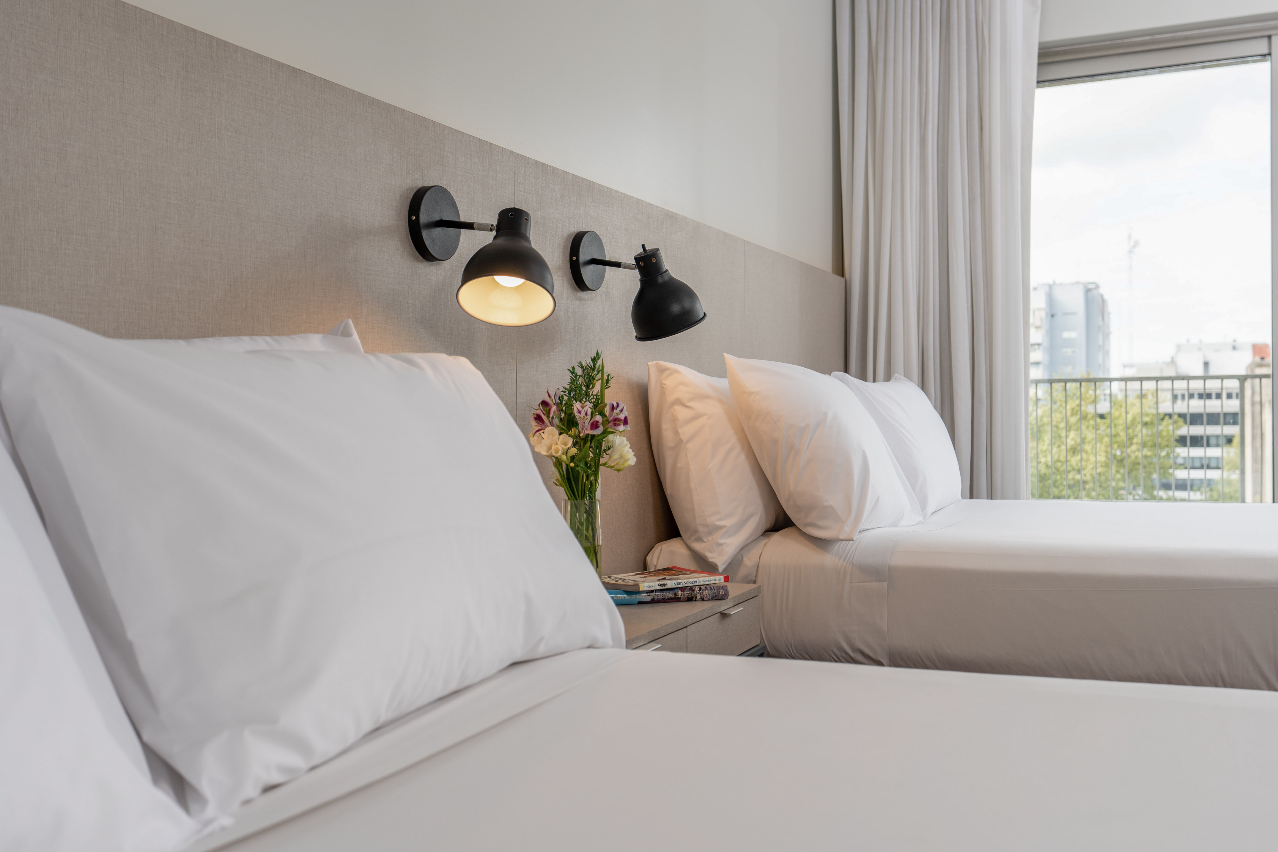 Hotel room with twin beds, white linens, modern wall lamps, and city view.