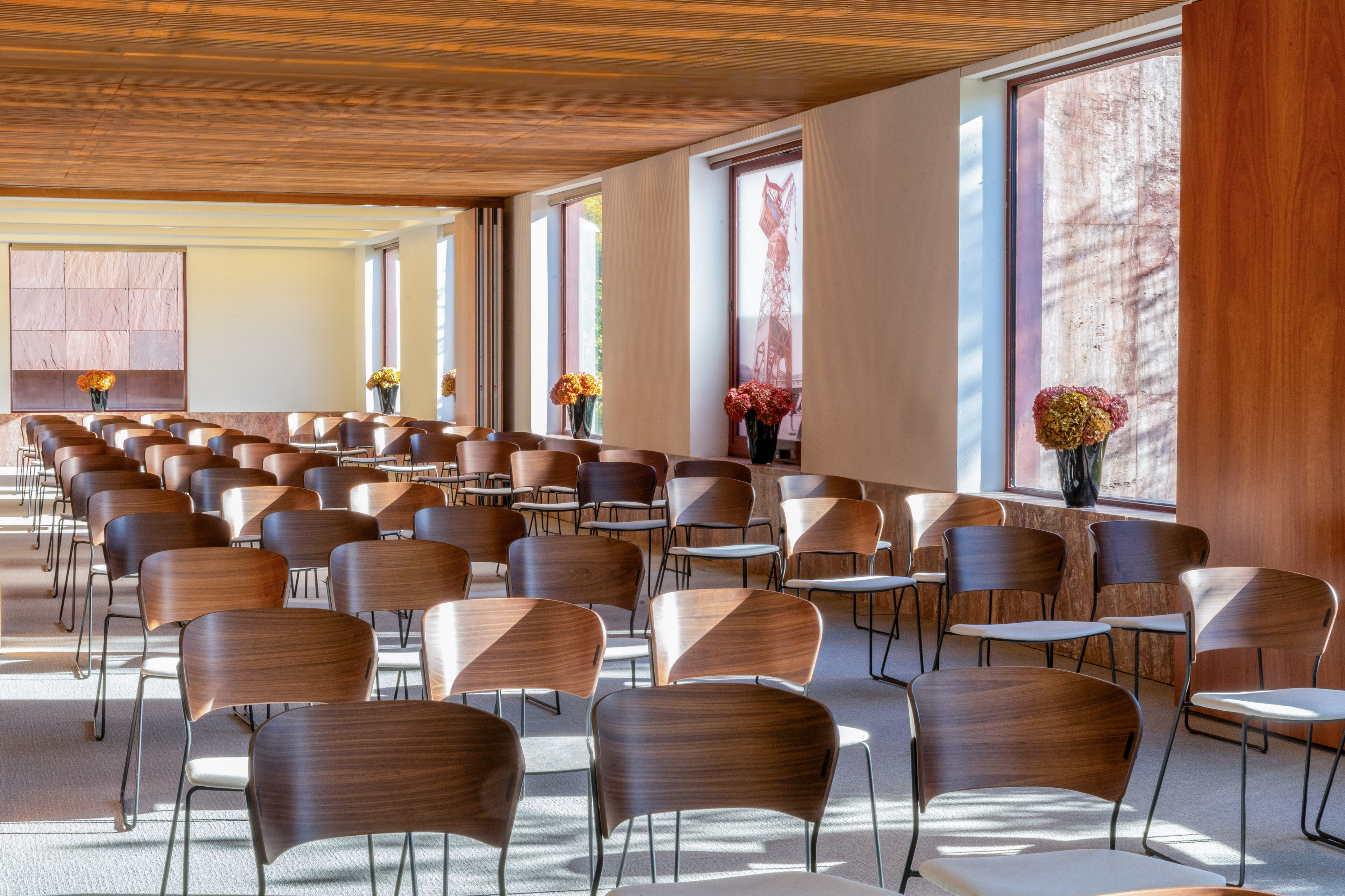 Modern lecture hall with wood chairs, sunlit windows, and wooden ceiling.