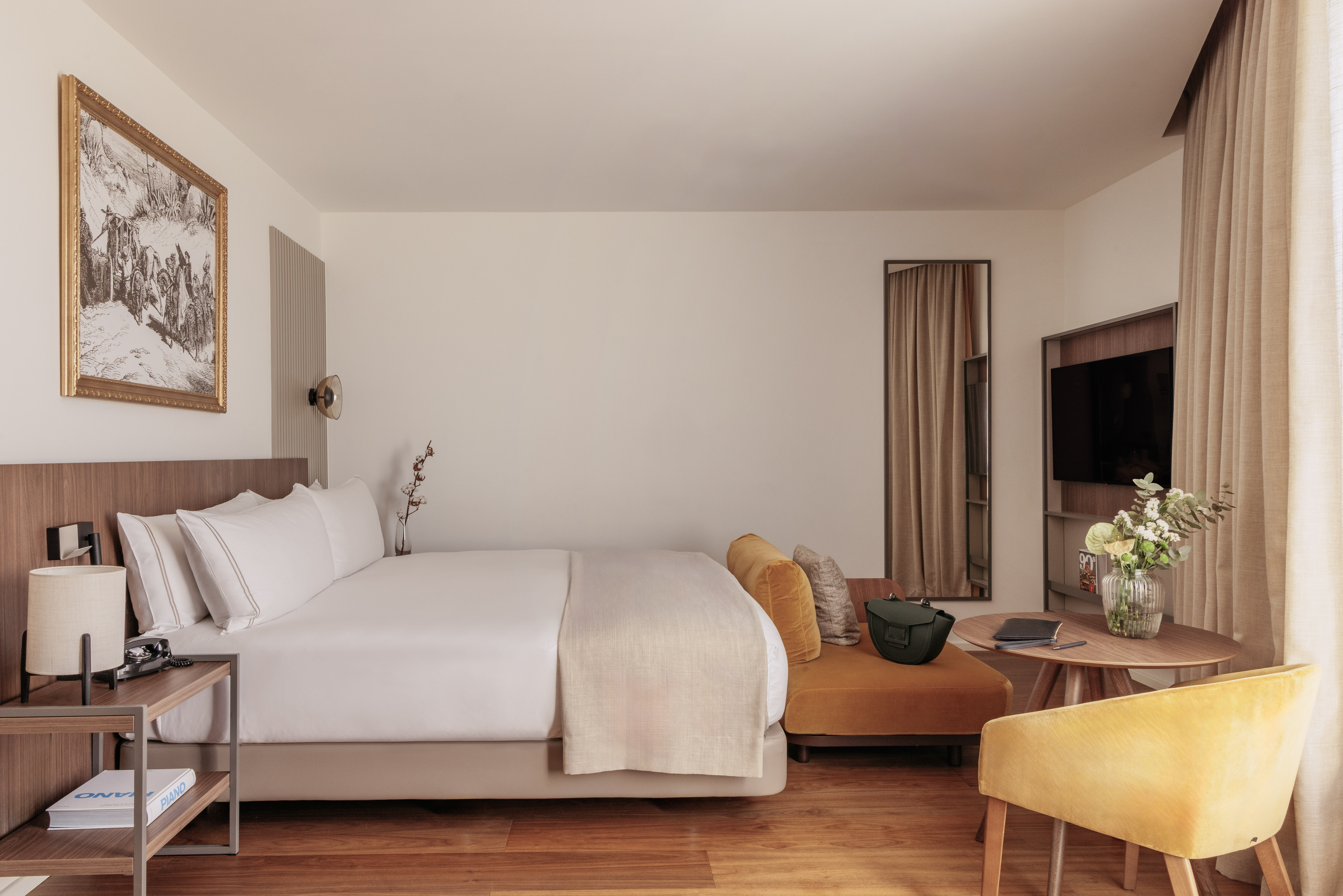 Modern hotel room with king bed, yellow sofa and armchair, wooden floor.