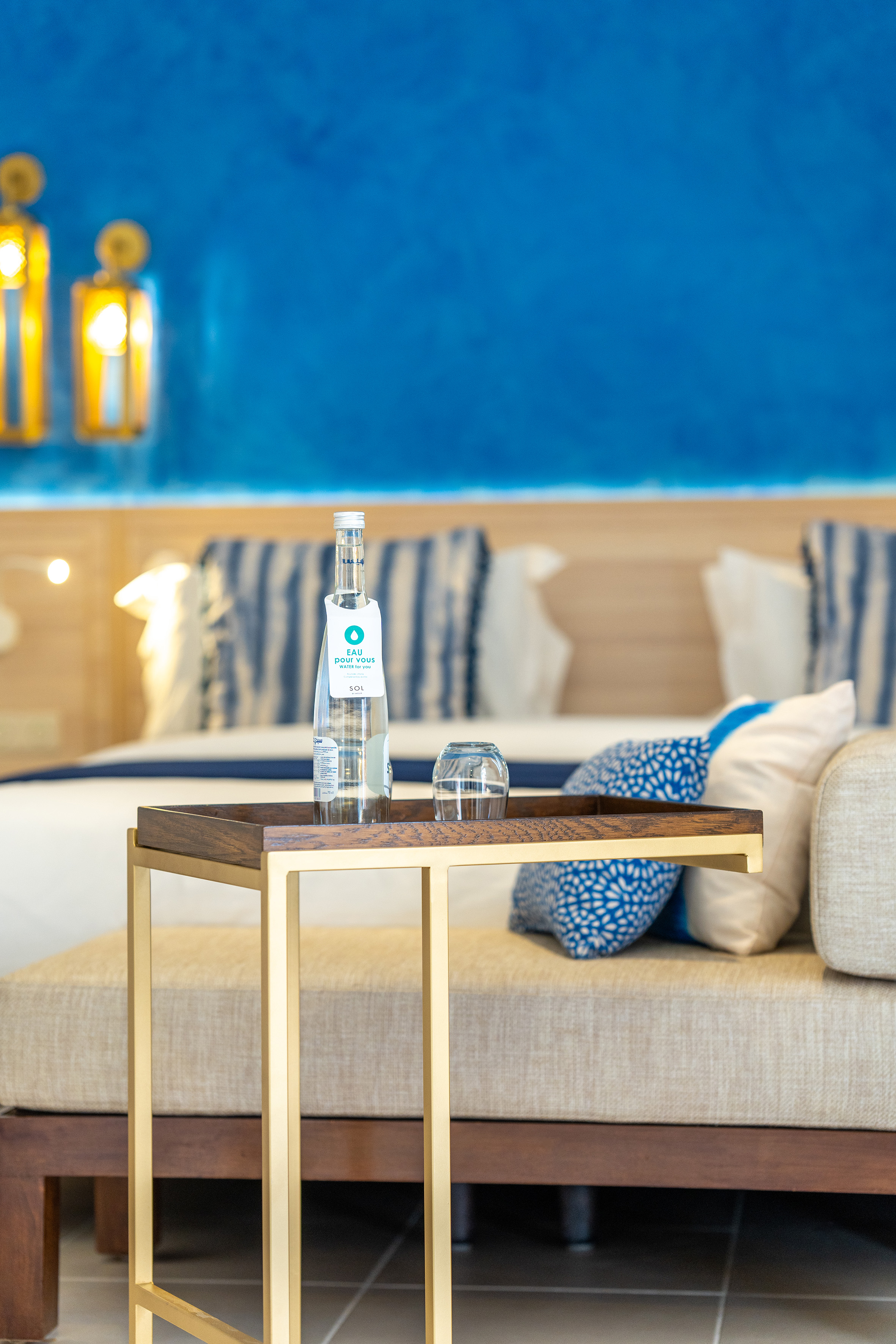 Water bottle & glass on modern gold side table in hotel room, blue wall.