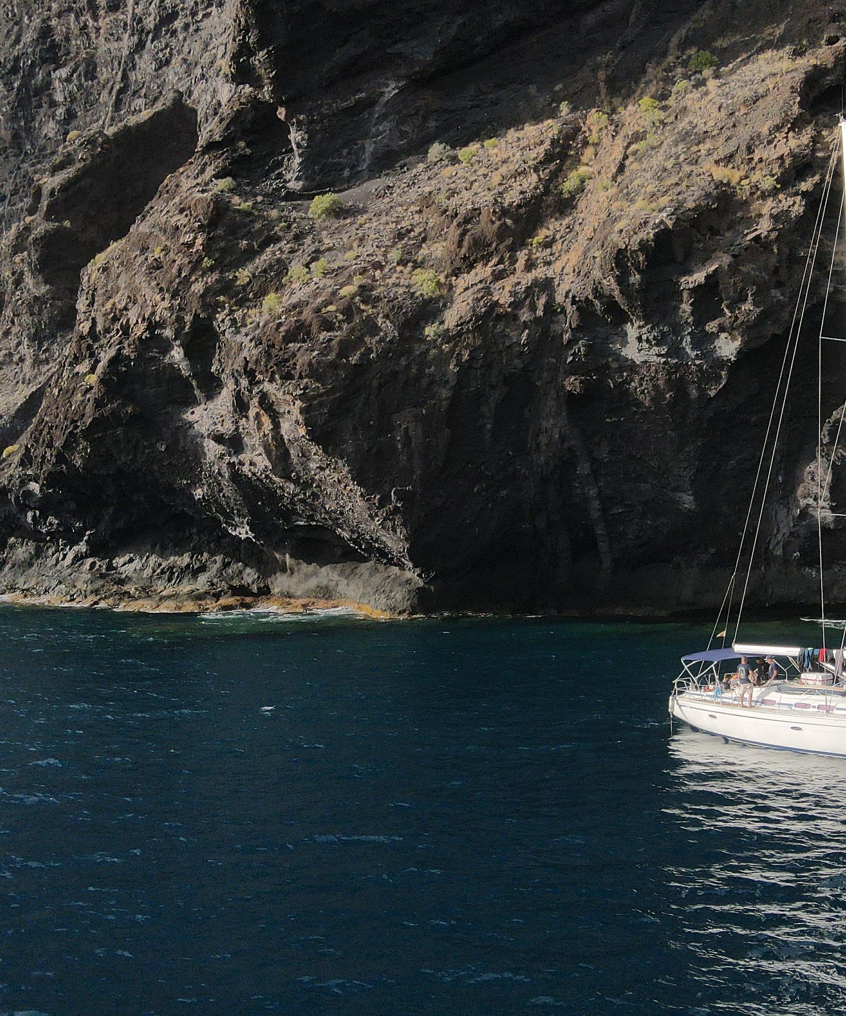 Sailboat anchored by a towering dark rocky cliff face in blue sea.
