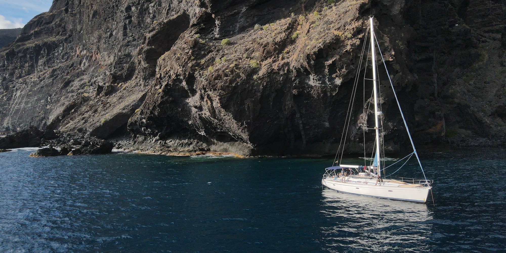 White sailboat on deep blue water by massive rugged cliffs.