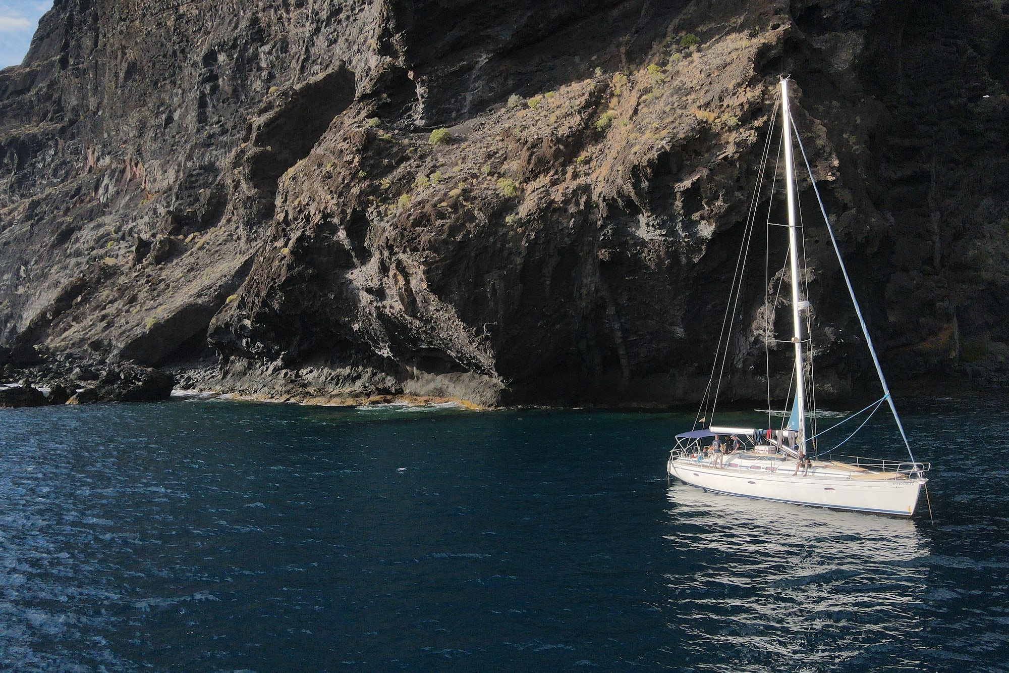 Sailboat anchored by a towering dark rocky cliff face in blue sea.