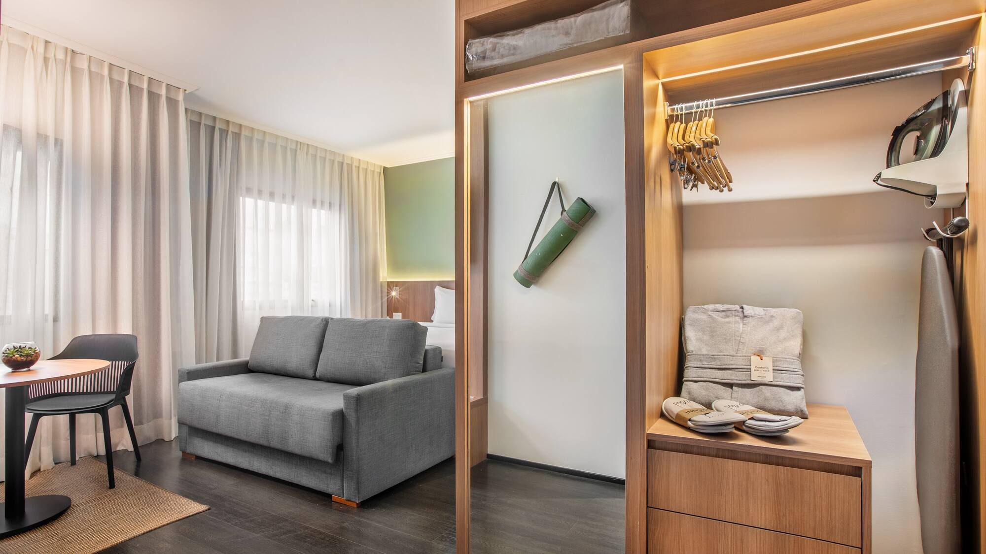 Modern hotel suite with sofa bed, wooden closet, yoga mat, and ironing board.