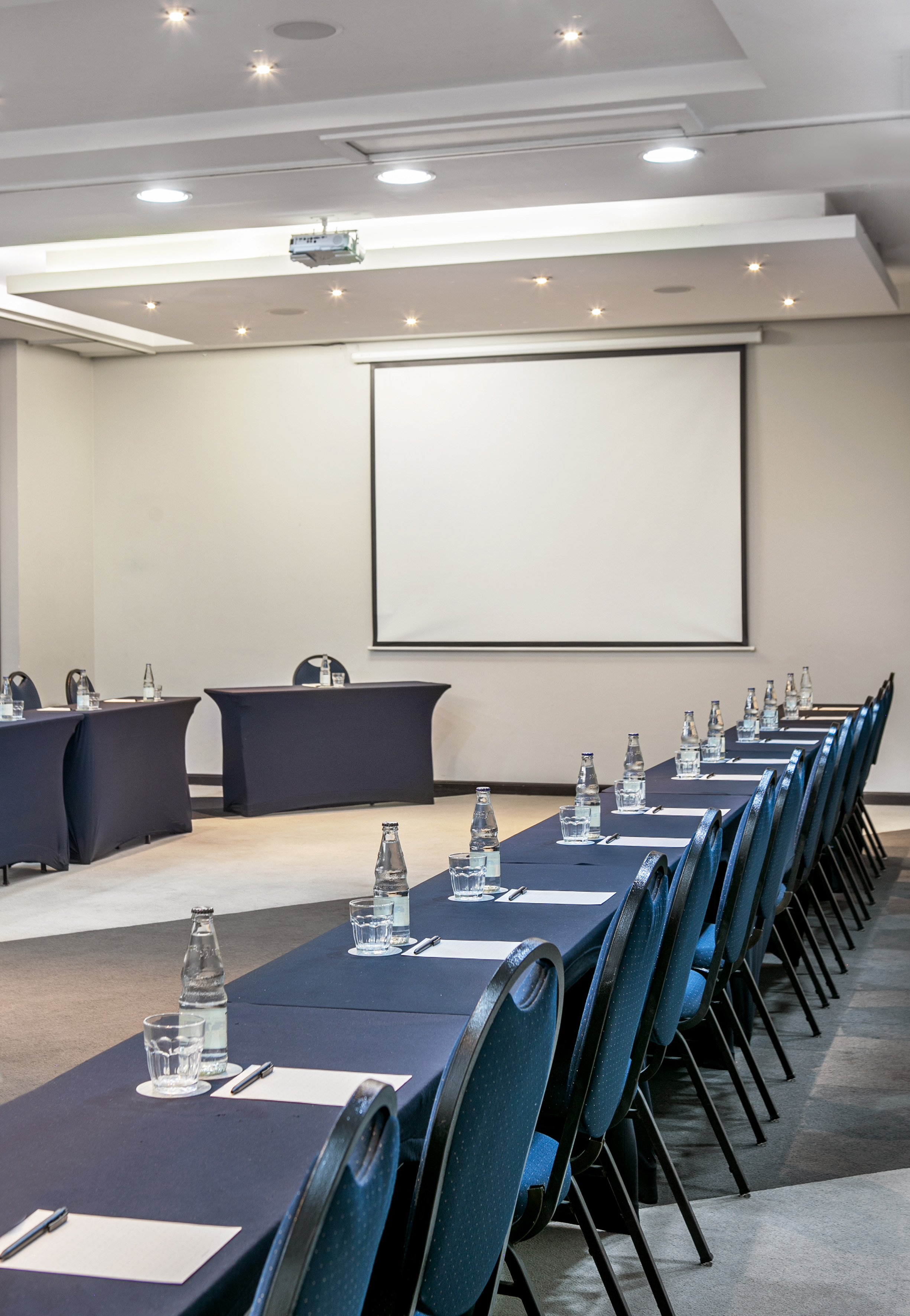 Empty conference room with blue chairs, tables, water, and projector screen.