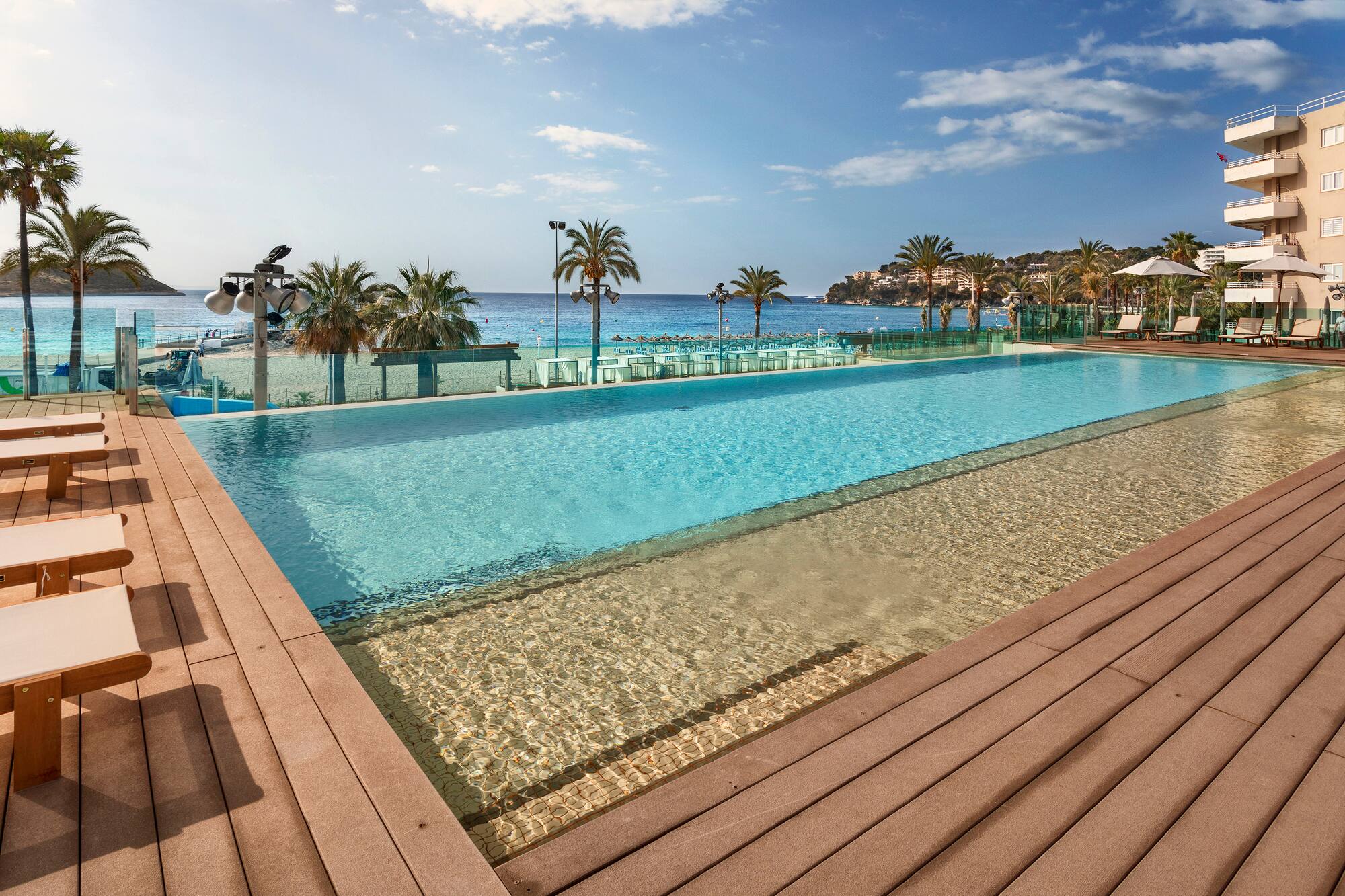 Resort infinity pool with ocean view, sandy beach, palm trees, and hotel.