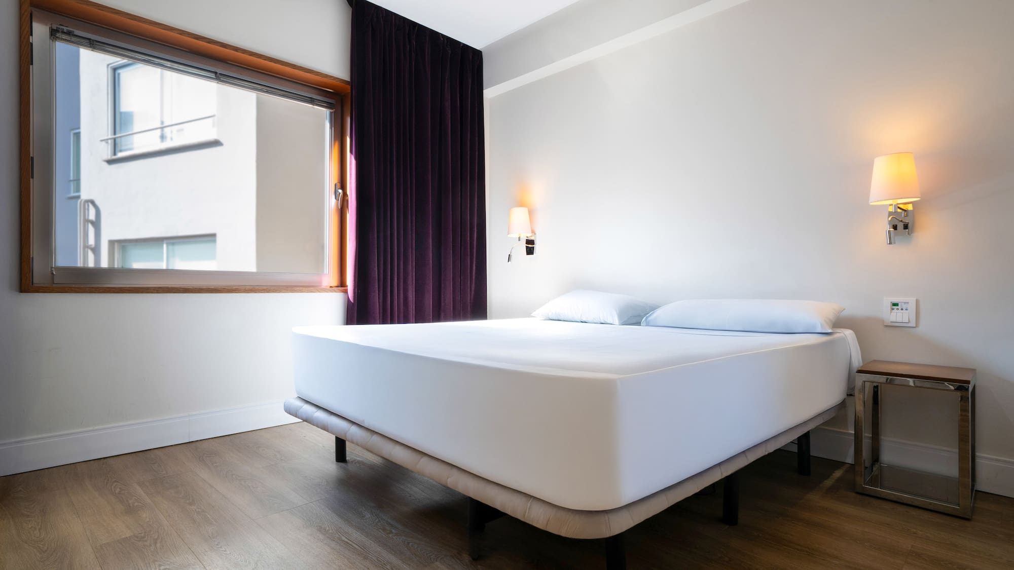 Hotel room with white bed, wooden floor, window, and purple curtains.