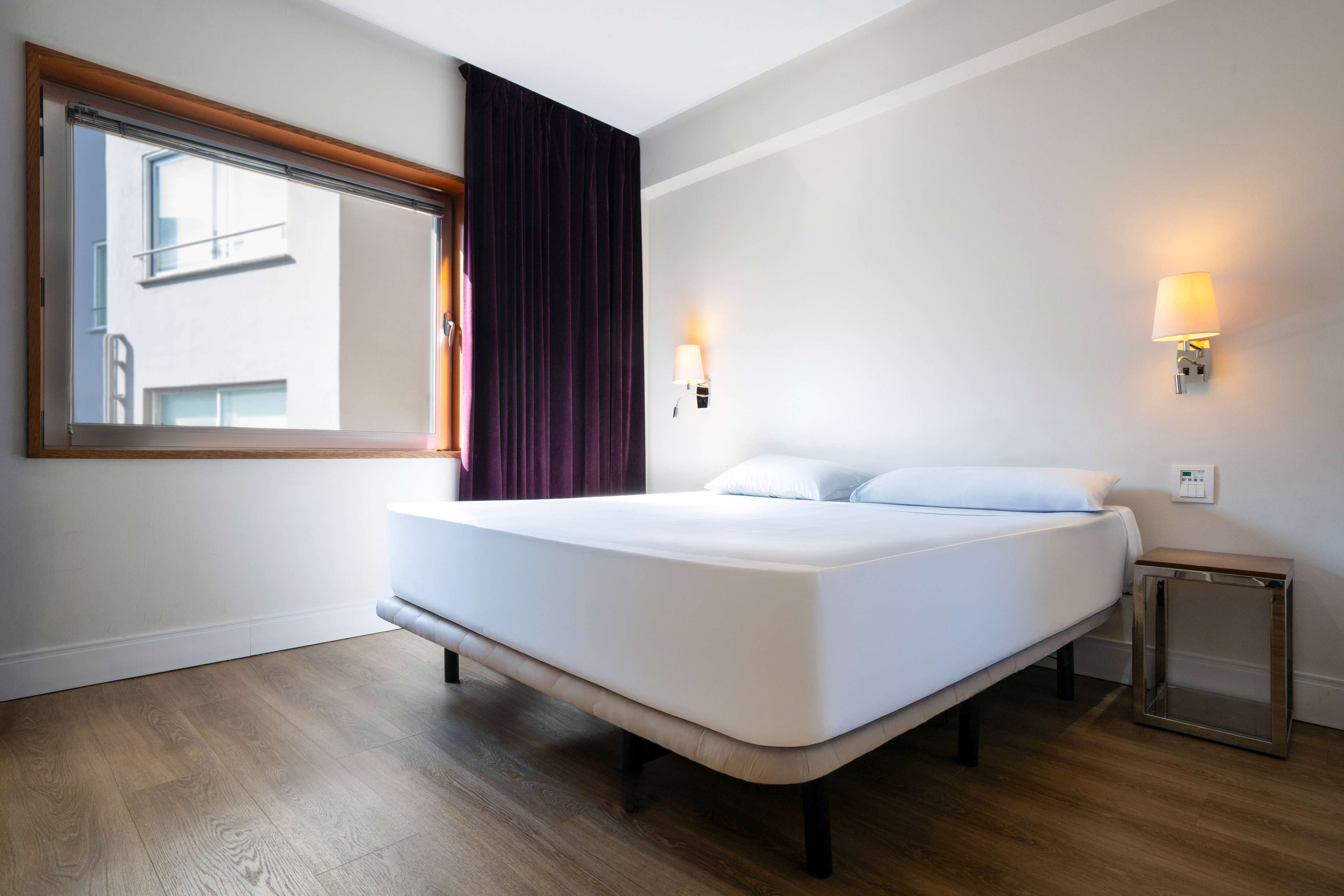 Hotel room with white bed, wooden floor, window, and purple curtains.