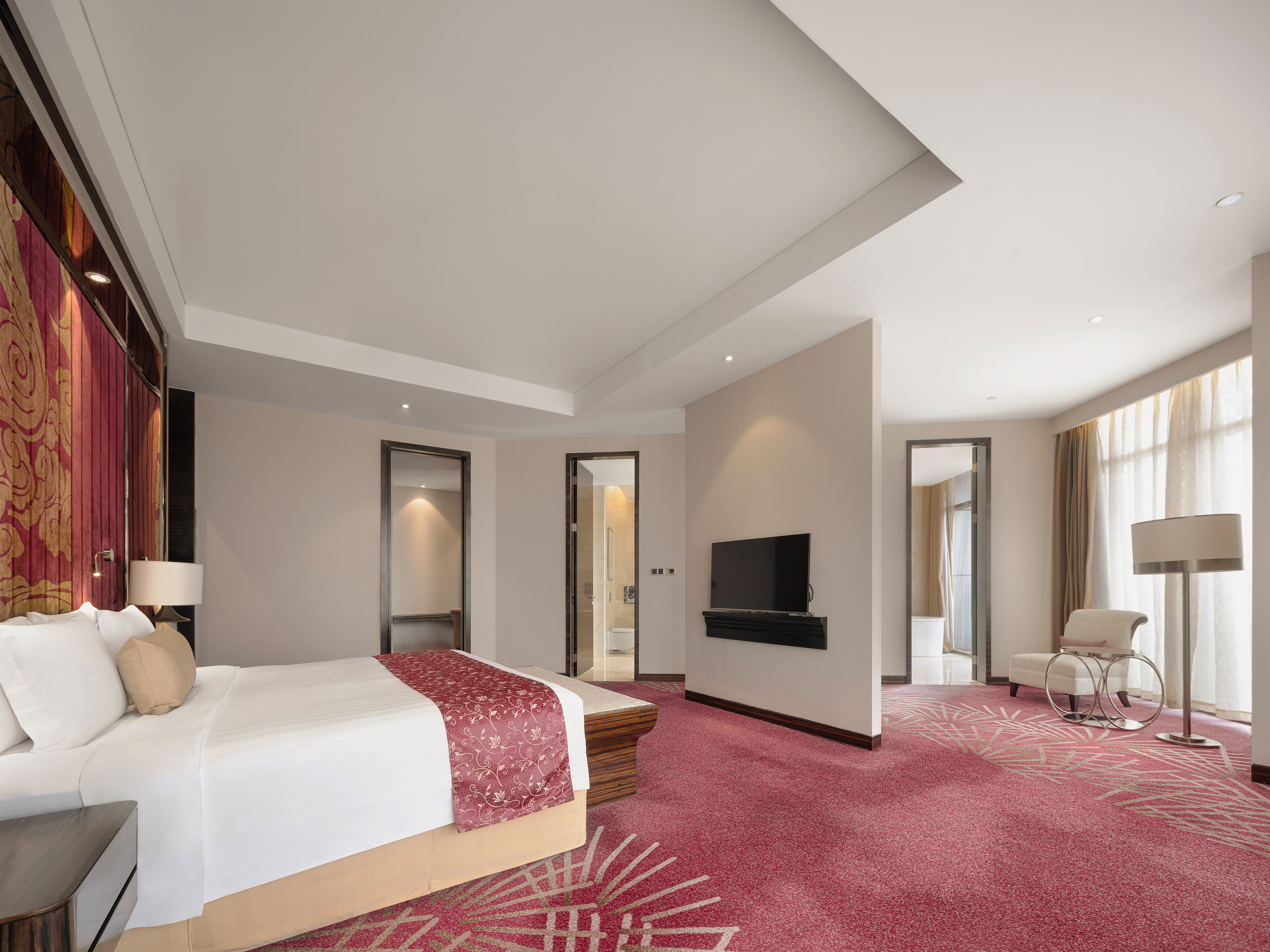 Spacious hotel bedroom suite with king bed, red patterned carpet, and TV.