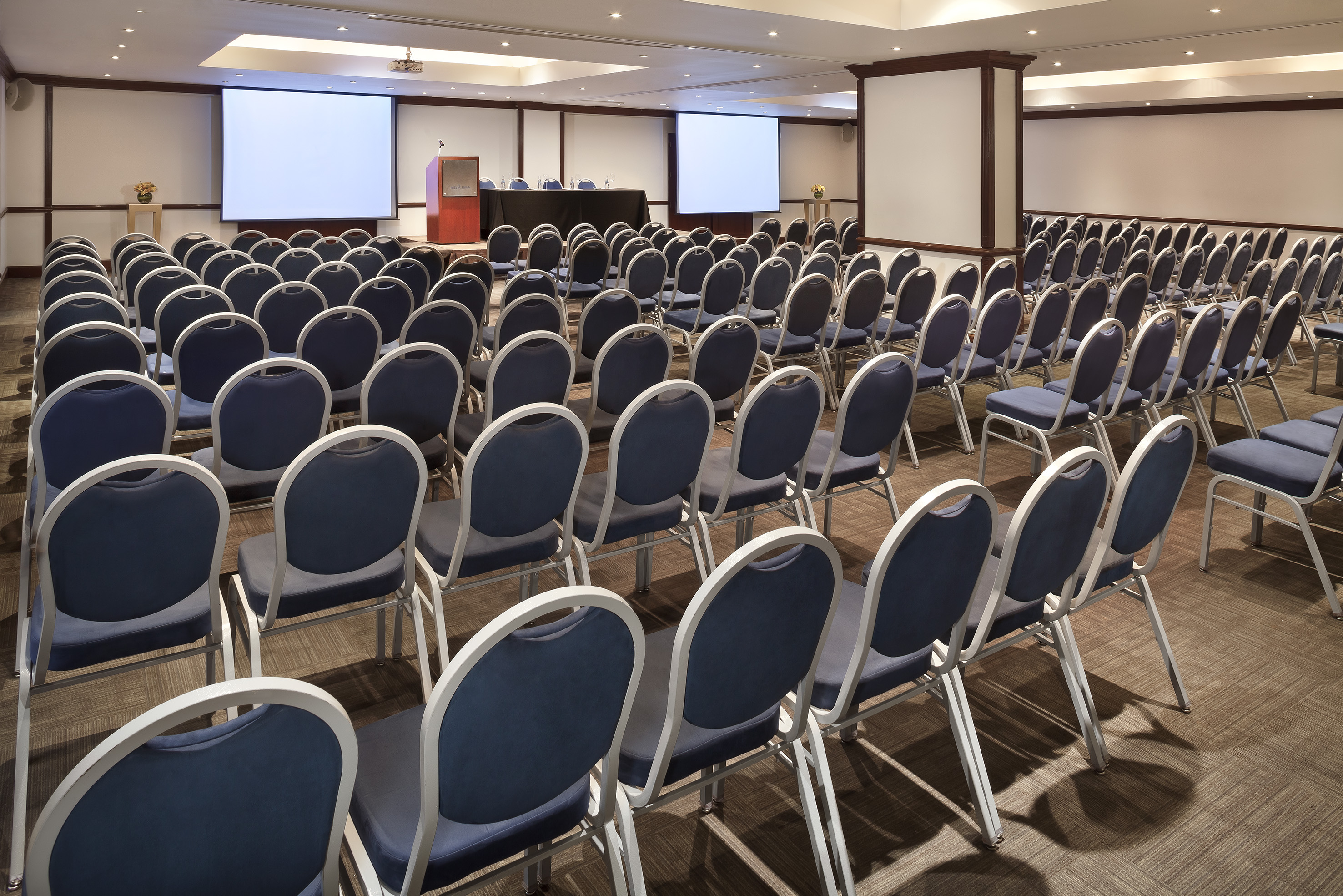 Spacious empty conference room setup with blue chairs, twin projection screens, and podium.