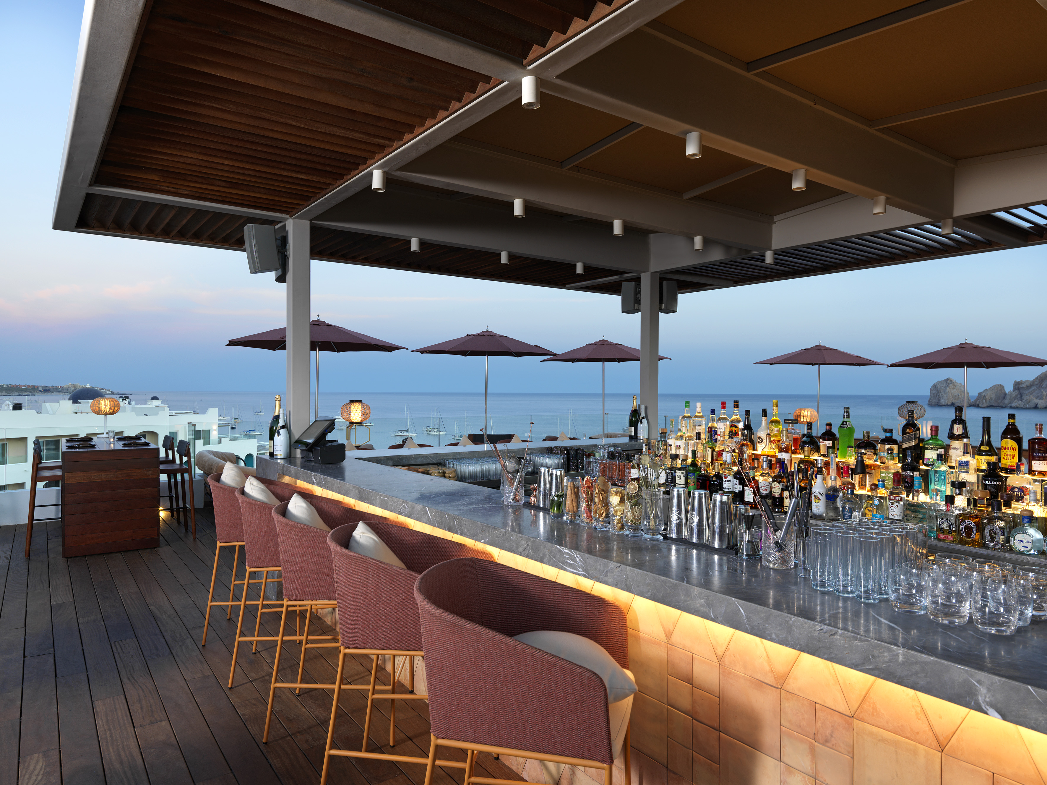 Rooftop bar with ocean view at dusk, liquor bottles, and red bar stools.