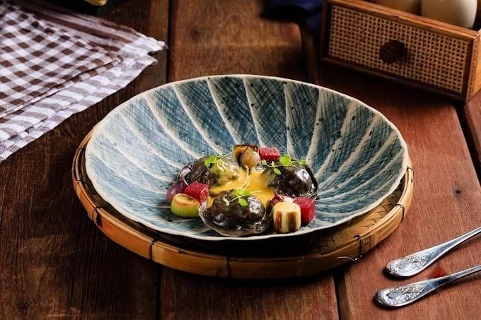 Abalone dish with yellow sauce, colorful garnishes in a patterned blue bowl on wood.