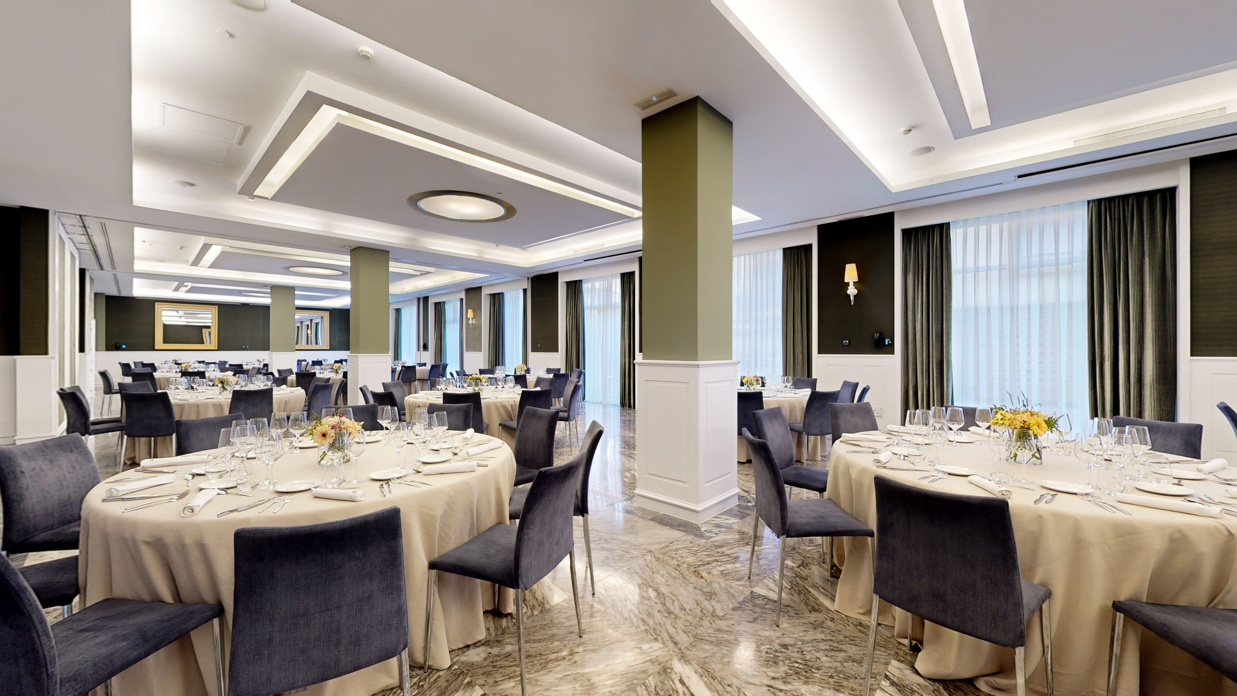 Modern event hall with round tables set for dining, velvet chairs, olive pillars.