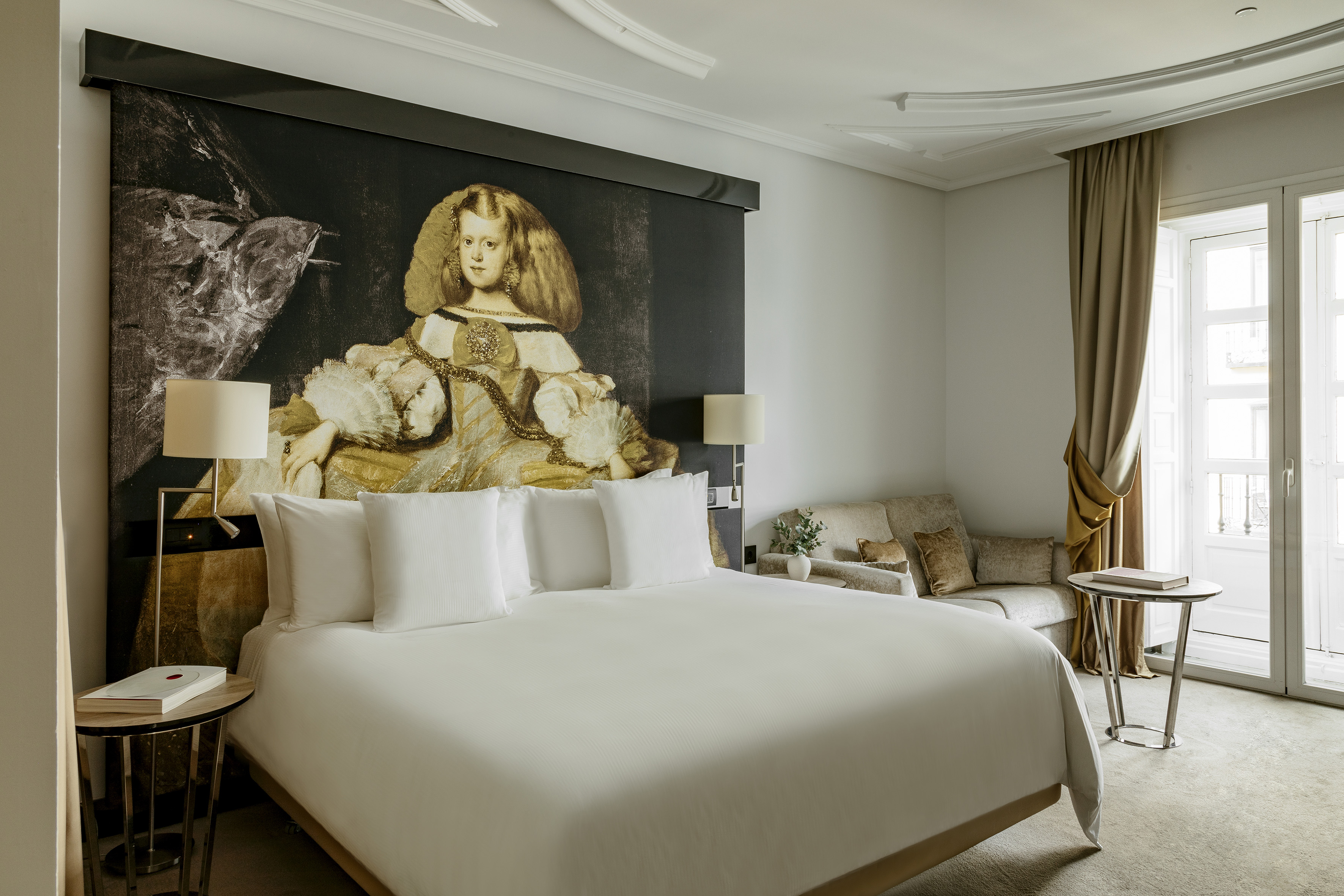 Luxury hotel bedroom with Las Meninas art wall, white bed, and elegant lounge.