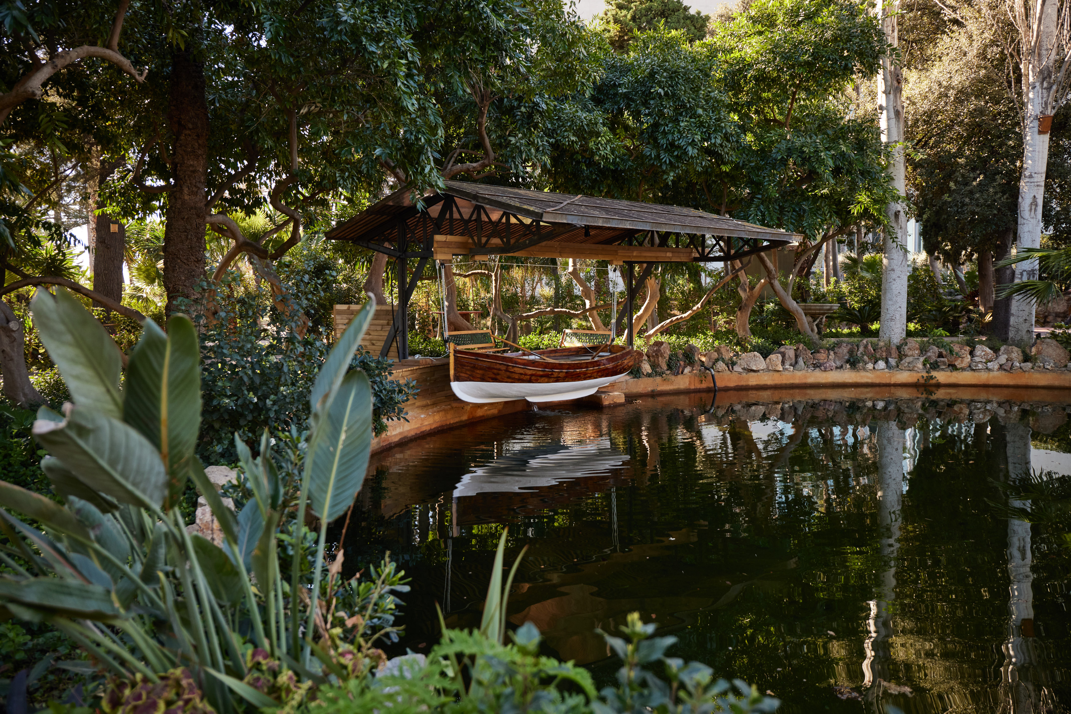 Wooden rowboat under a covered shelter, over a pond in a lush garden.