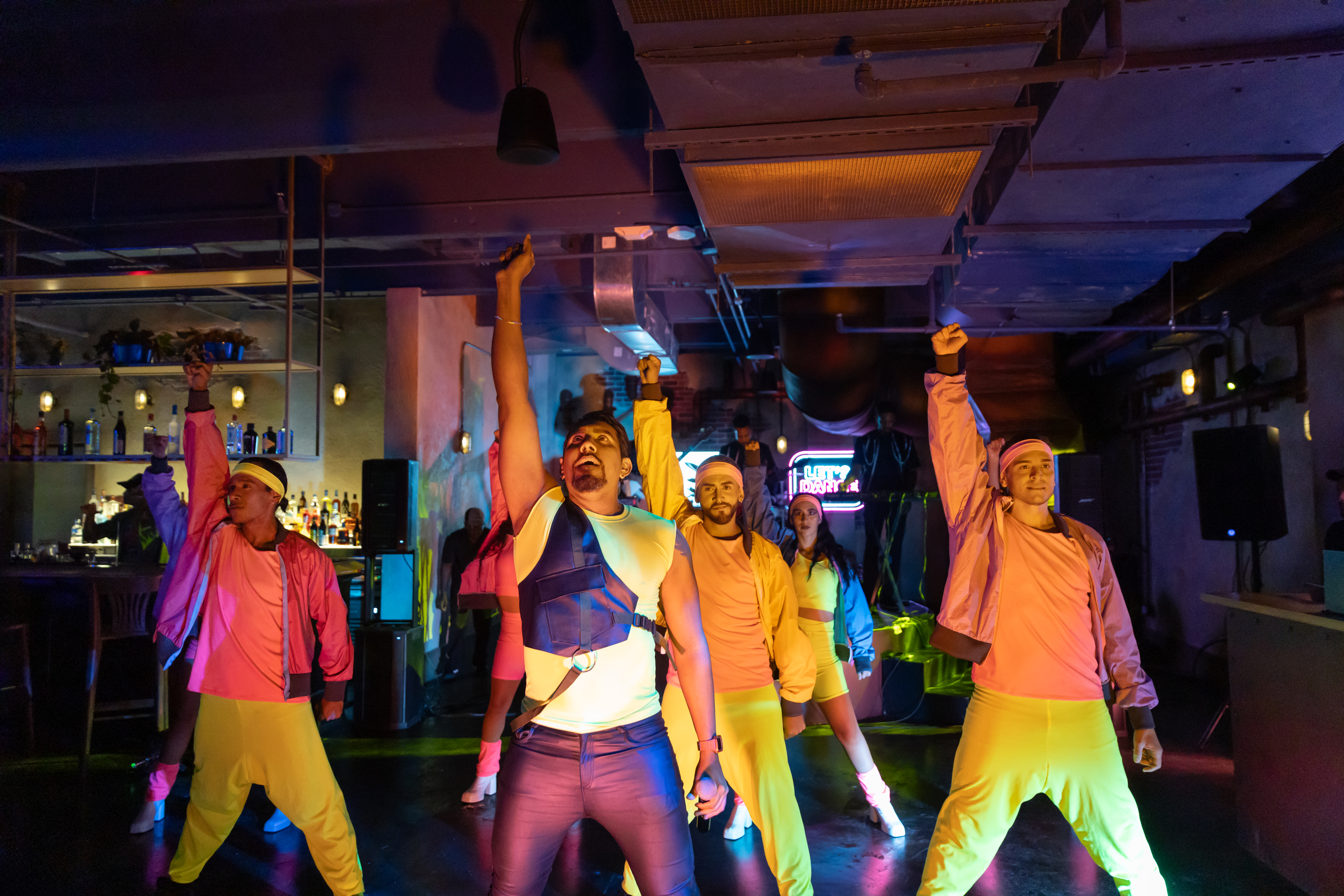Dancers in bright neon 80s attire raising arms under colorful stage lights.