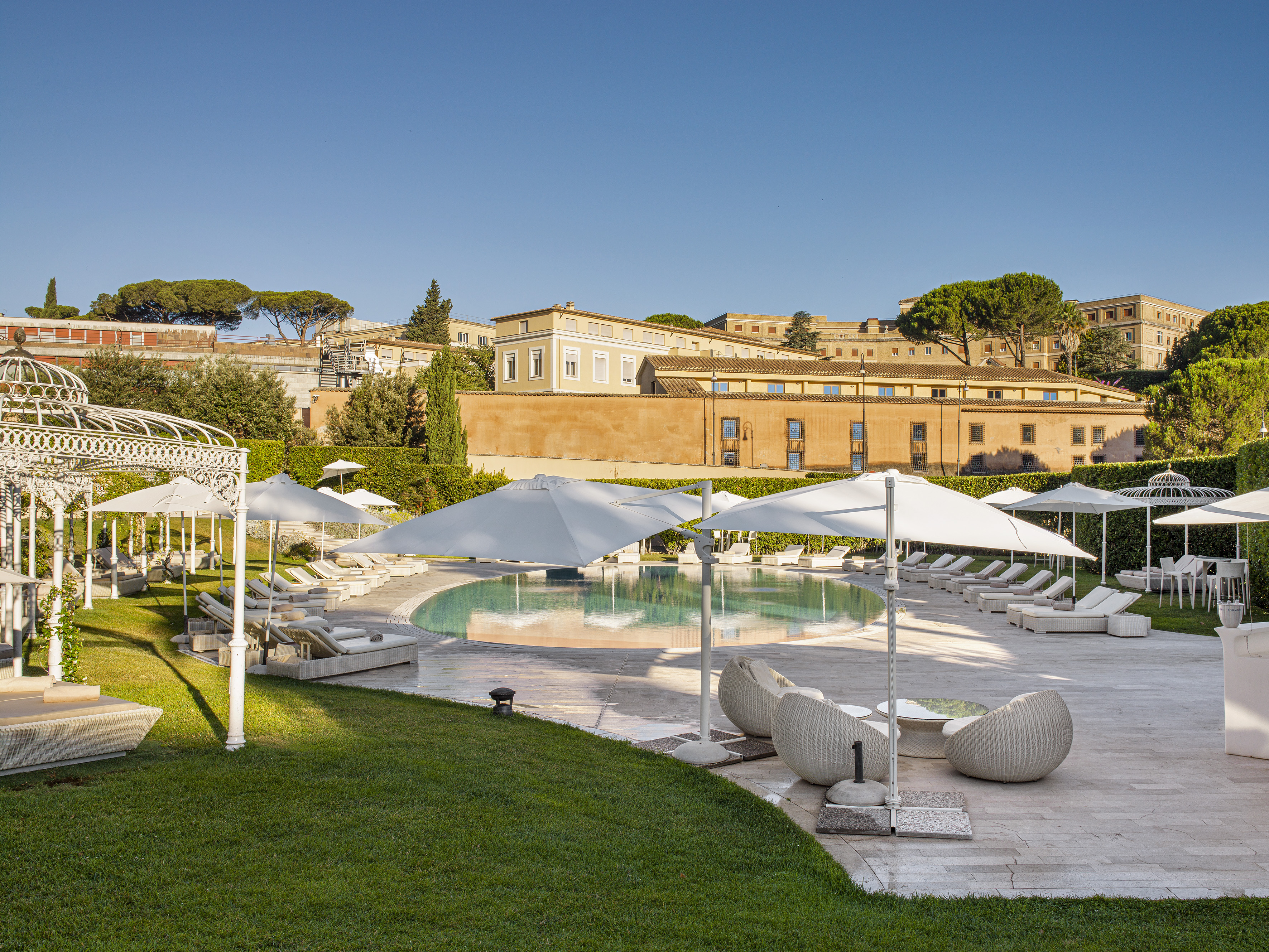 Sunny resort pool with lounge chairs, umbrellas, green lawn, and buildings.