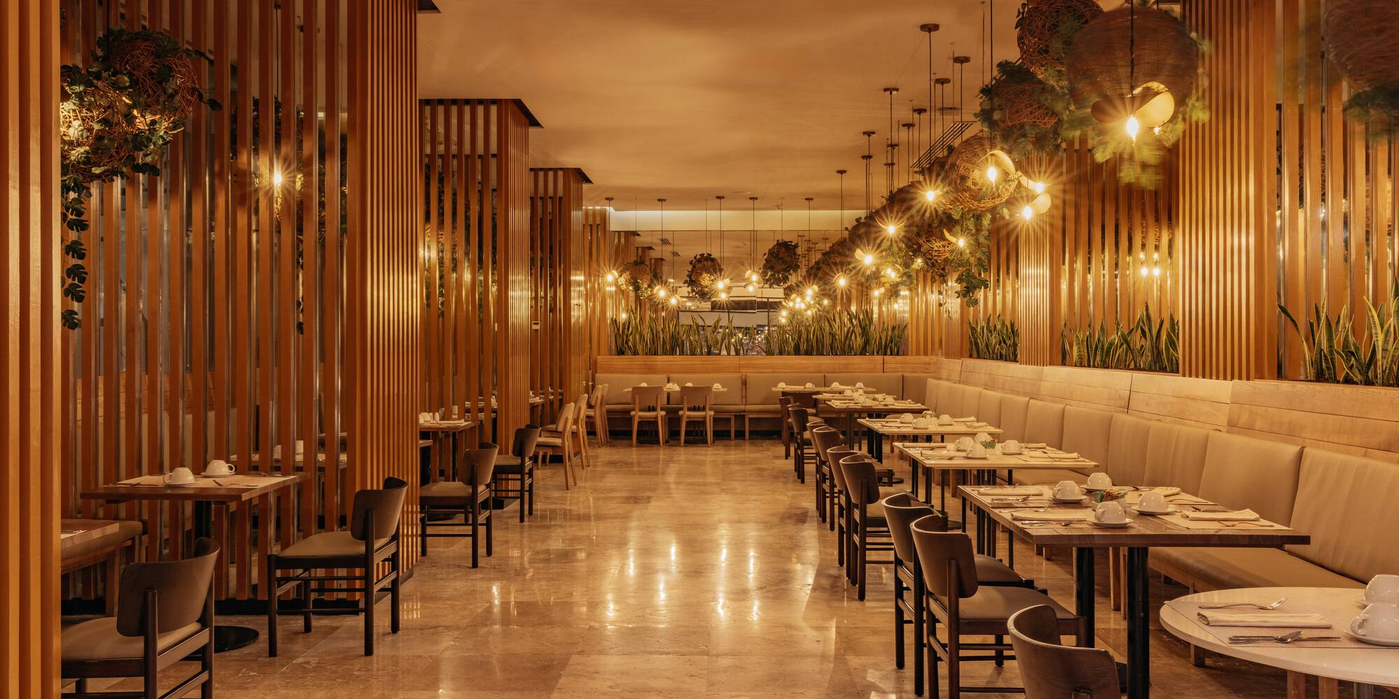 Warm-lit restaurant interior with wooden slats, lush plants, and set dining tables.