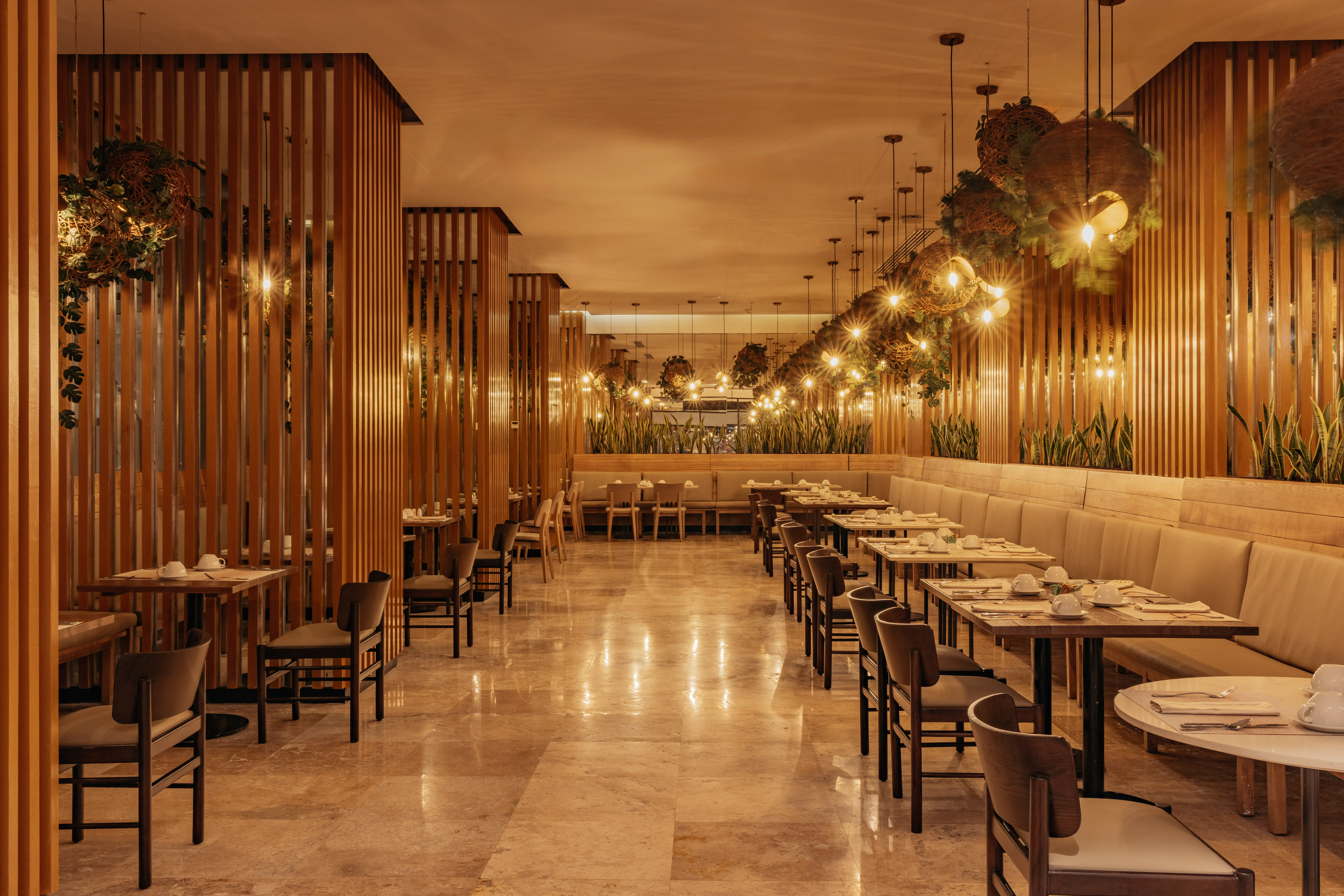 Warm-lit restaurant interior with wooden slats, lush plants, and set dining tables.