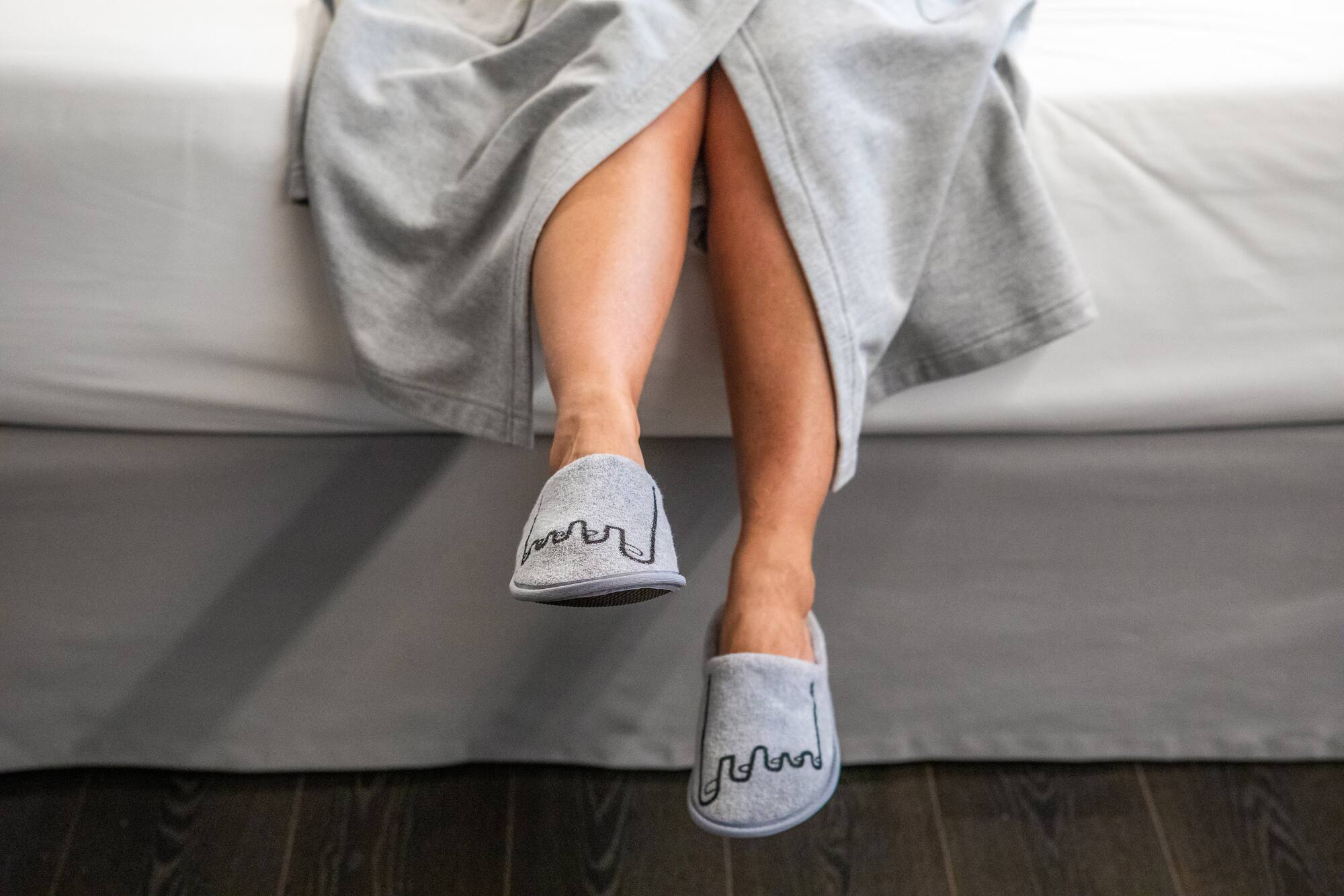 Legs in gray robe and gray embroidered slippers at bed edge.