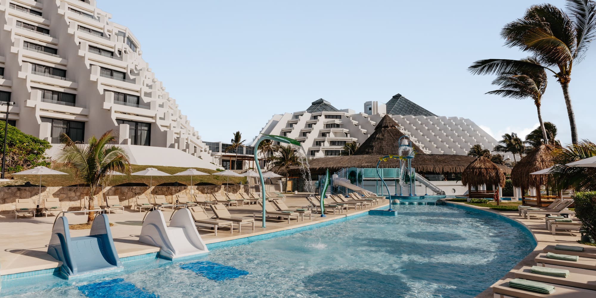 Sunny resort pool with waterslides, palm trees, and pyramid-style hotel buildings.
