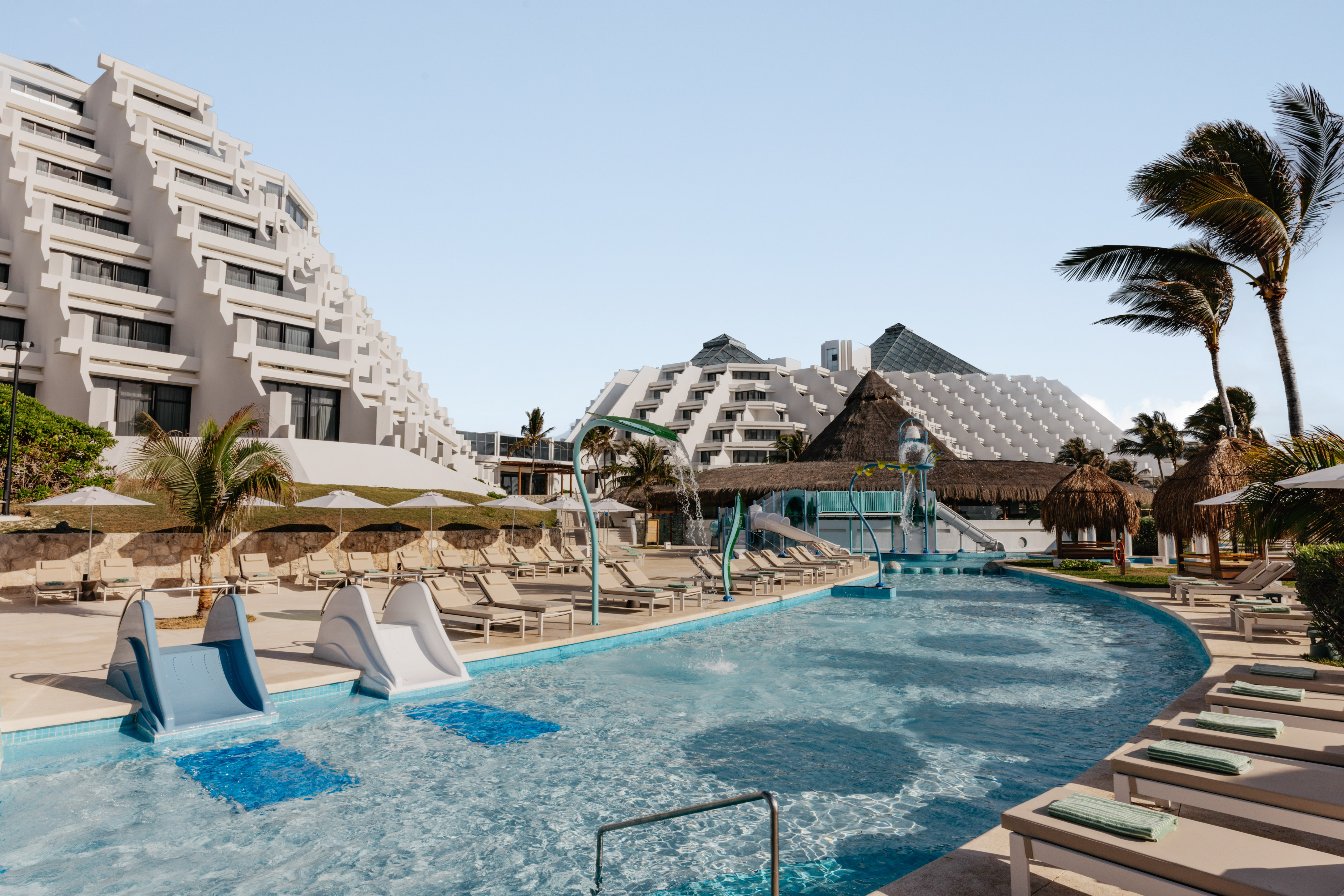 Sunny resort pool with waterslides, palm trees, and pyramid-style hotel buildings.