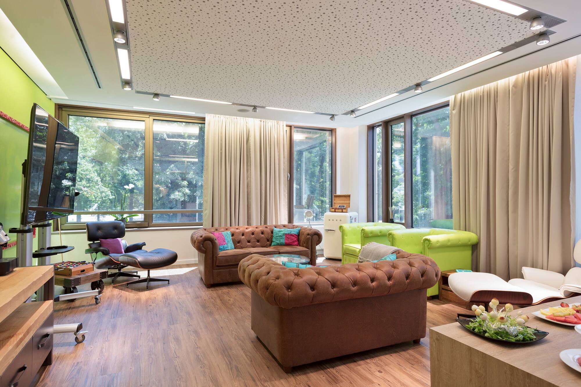 Modern lounge with brown leather sofas, green armchairs, large windows, wood floor.