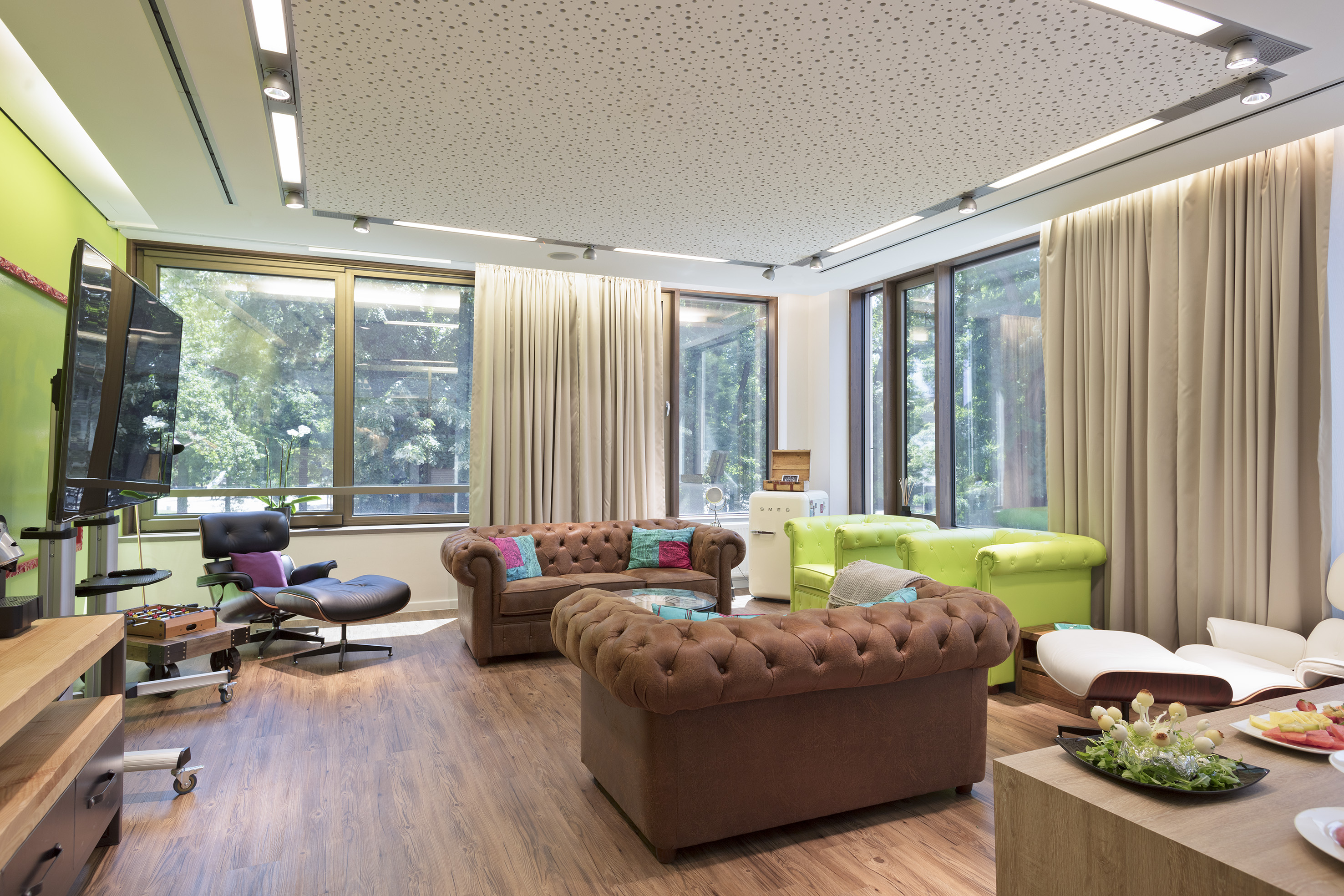 Modern lounge with brown leather sofas, green armchairs, large windows, wood floor.