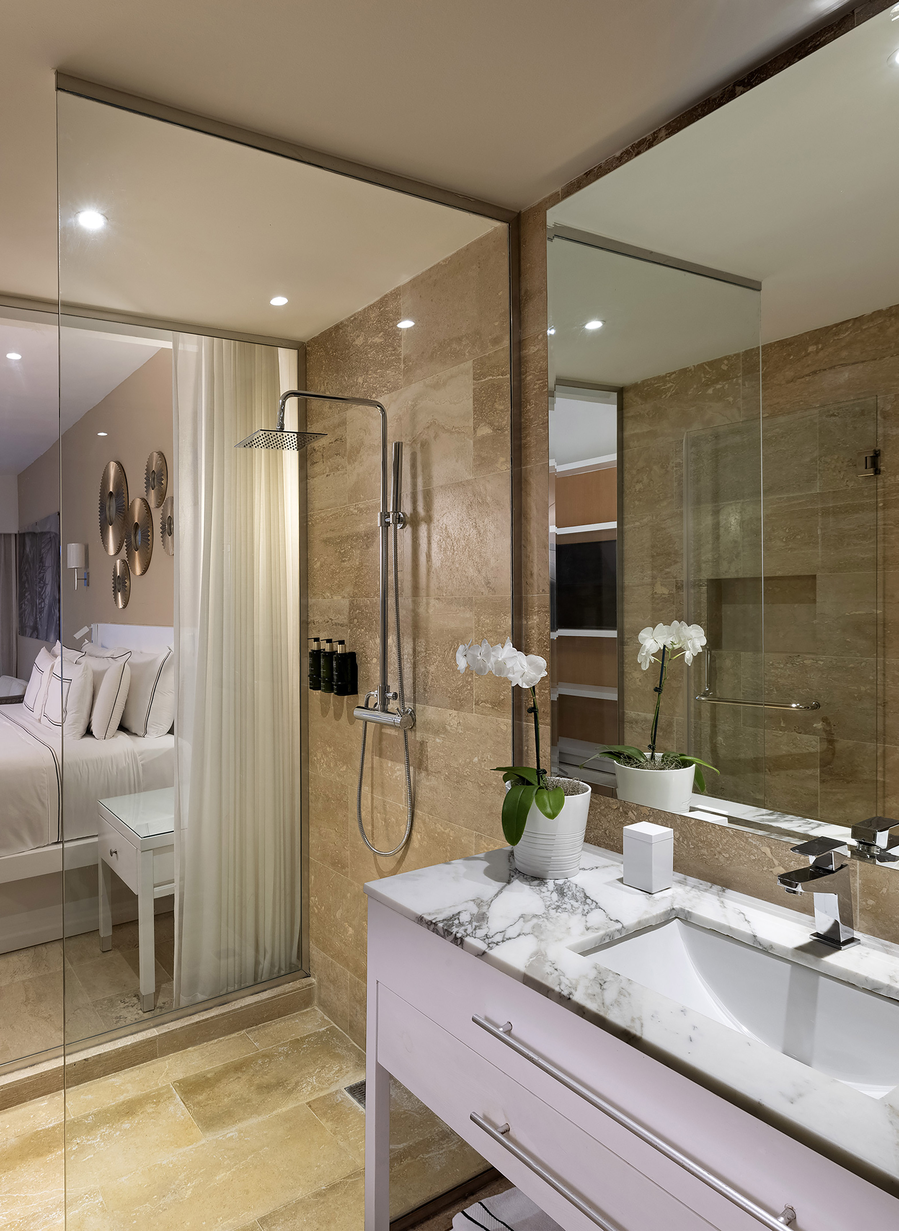 Modern bathroom with walk-in shower, marble vanity, and bedroom visible through glass.