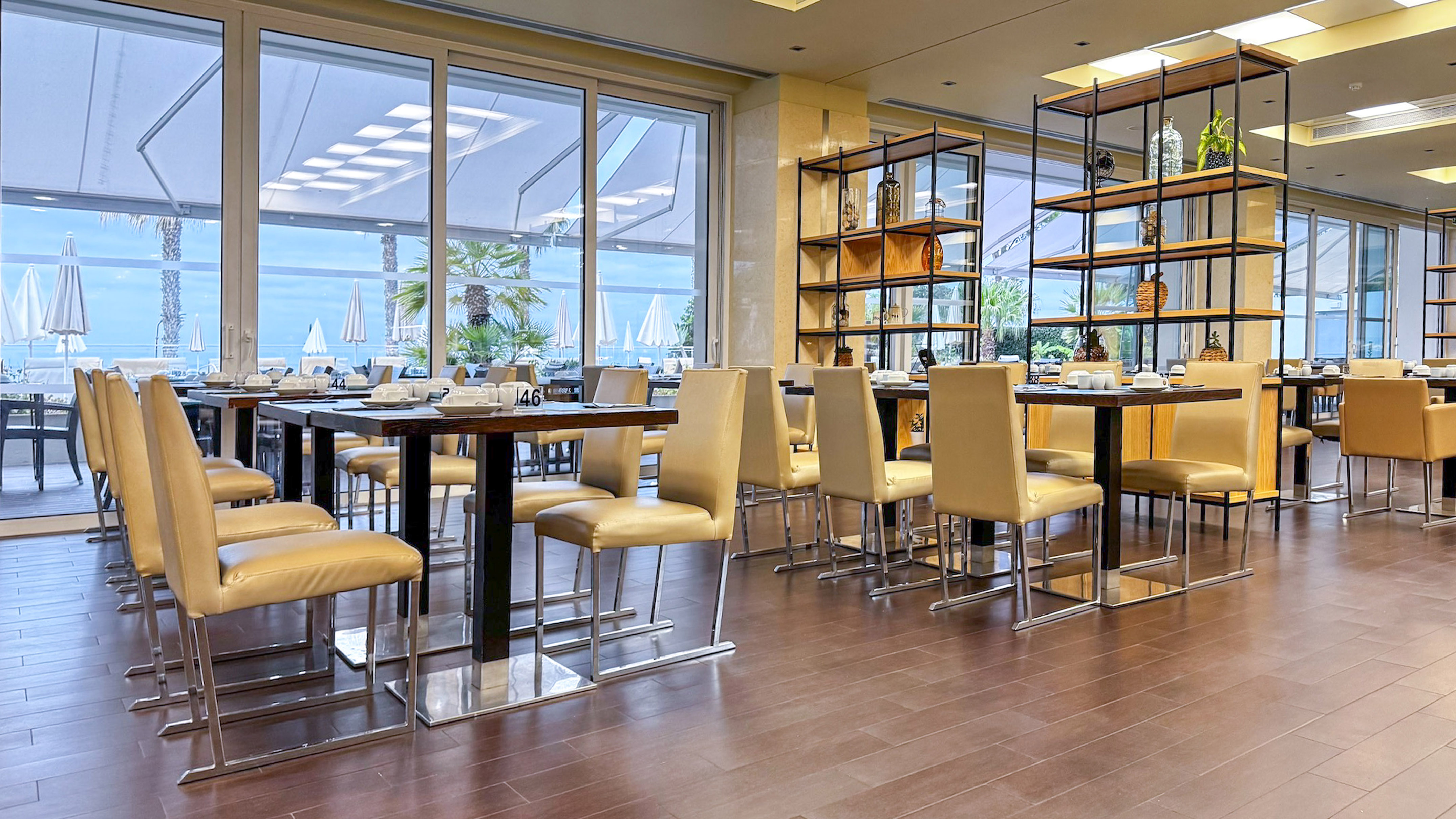 Modern restaurant with sea view, set dining tables, and beige chairs.