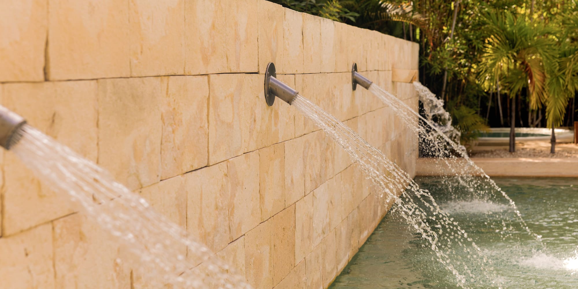 Stone wall water spouts filling a pool in a tropical setting.