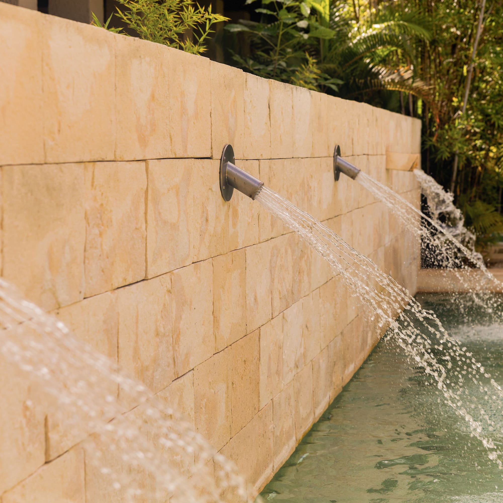 Stone wall water spouts filling a pool in a tropical setting.