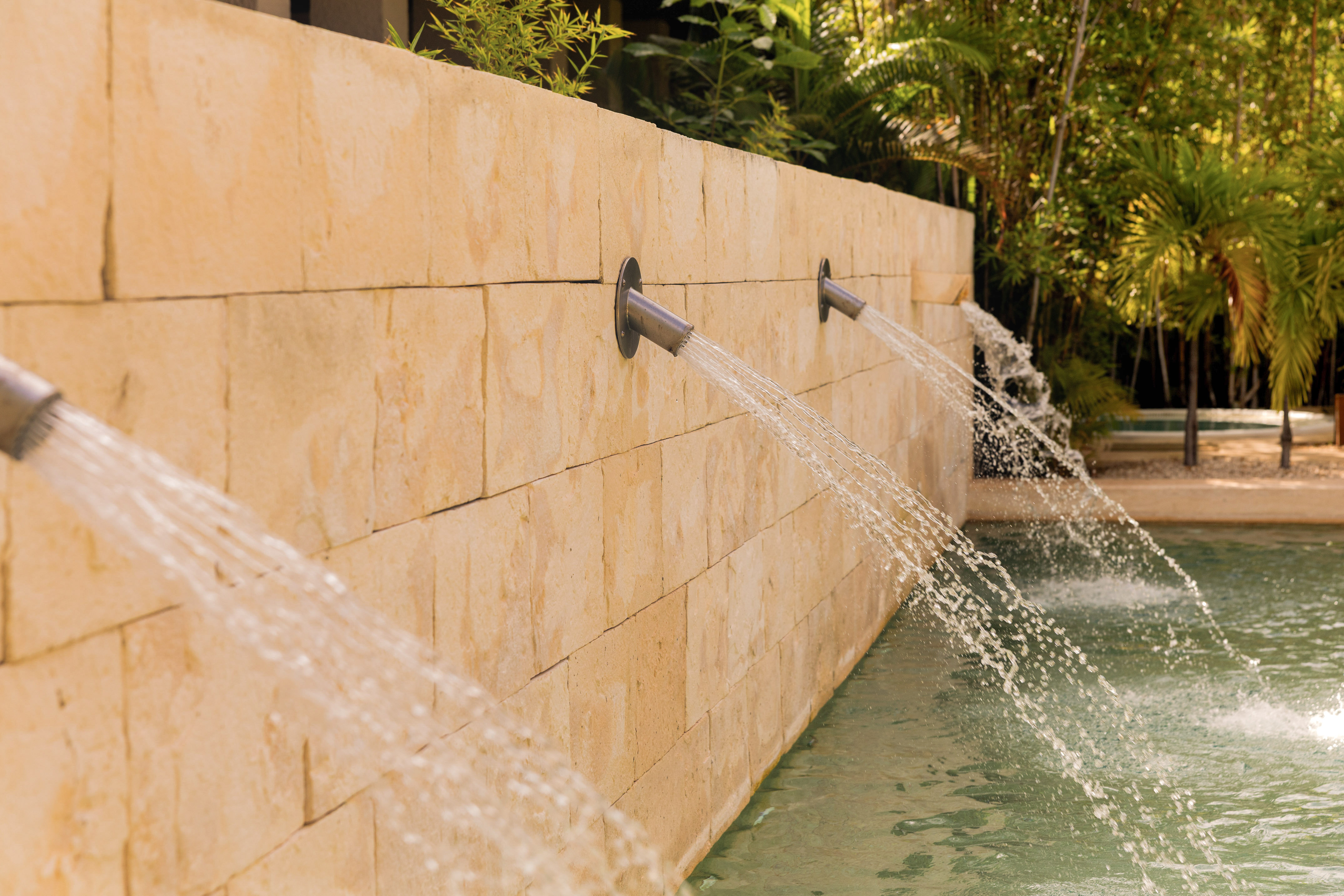 Stone wall water spouts filling a pool in a tropical setting.