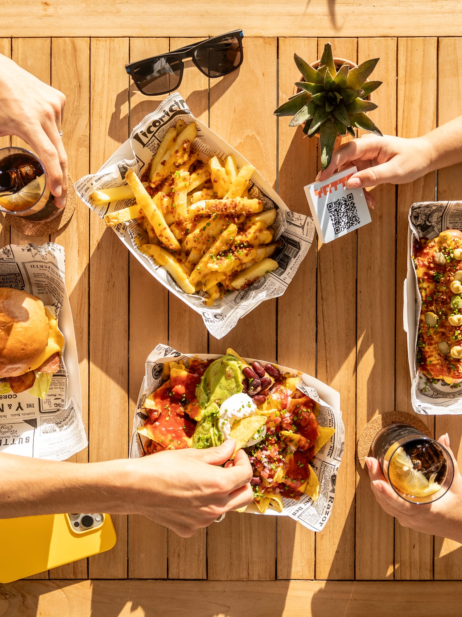 Friends sharing burgers, fries, nachos, hot dog, drinks on outdoor table.