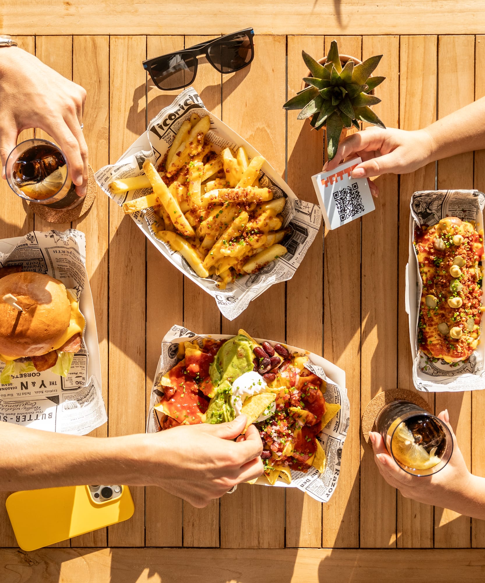 Hands reaching for burger, fries, nachos, hot dog, drinks on wooden table.