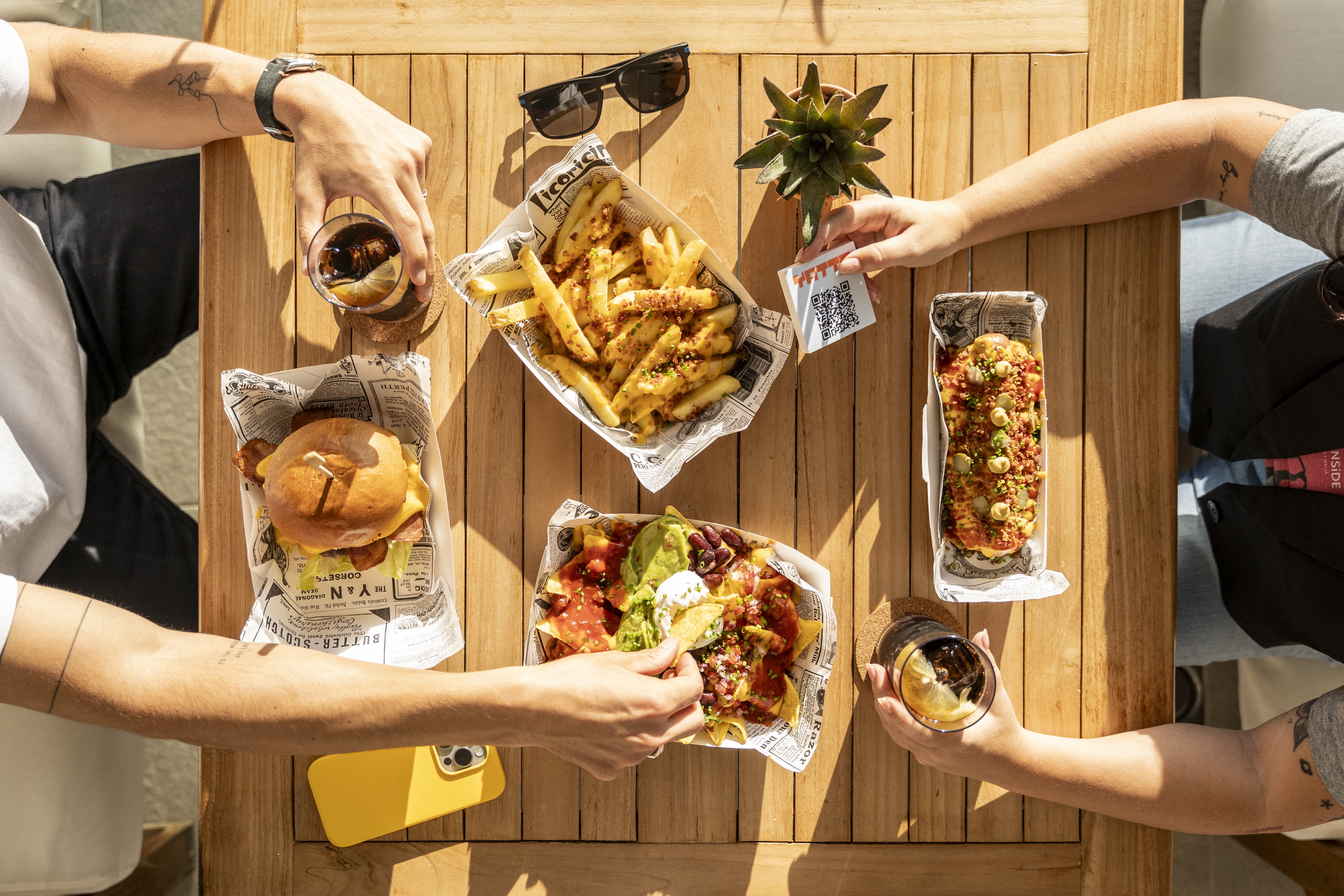 Hands reaching for burger, fries, nachos, hot dog, drinks on wooden table.