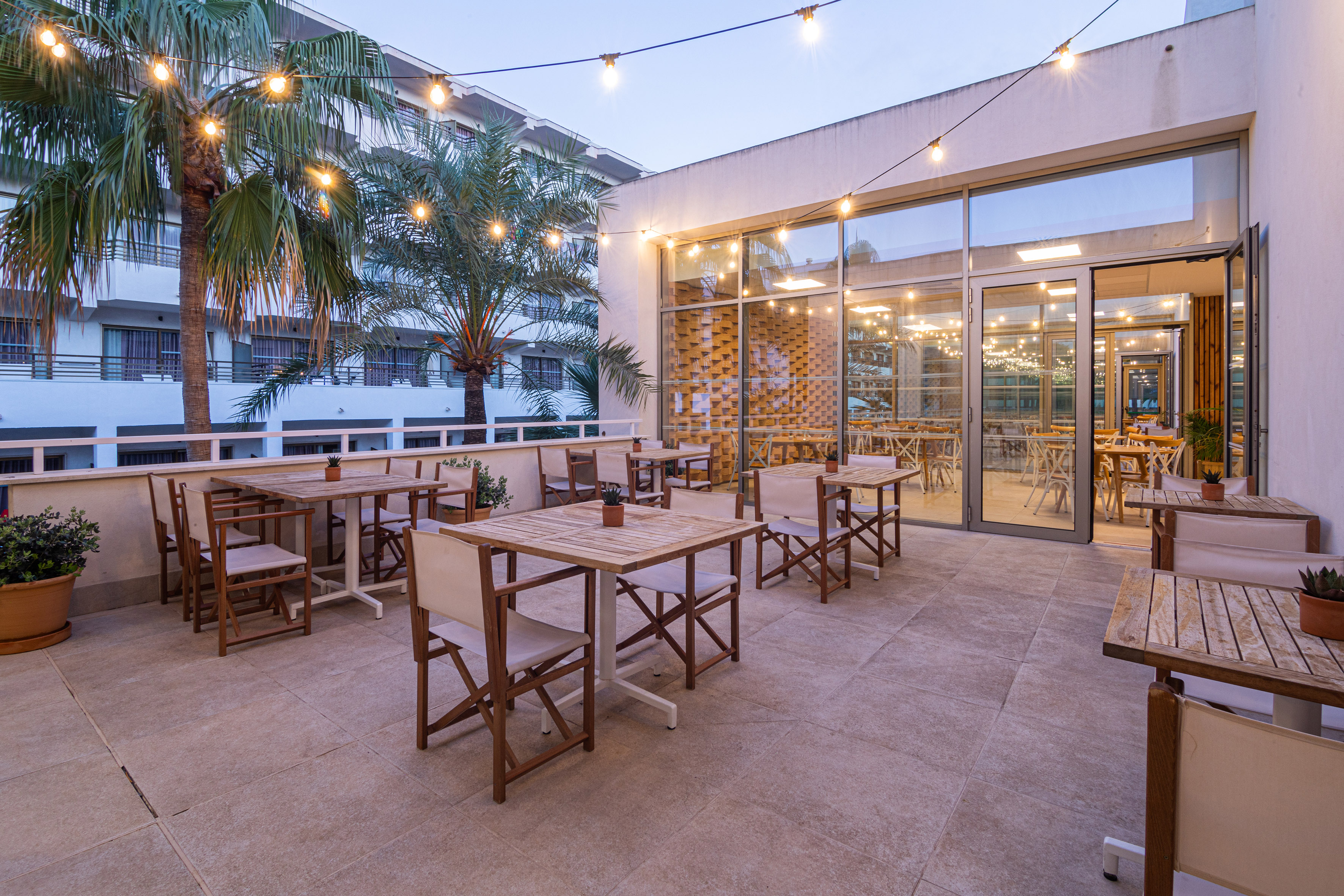 Resort outdoor patio with dining tables, string lights, and palm trees.
