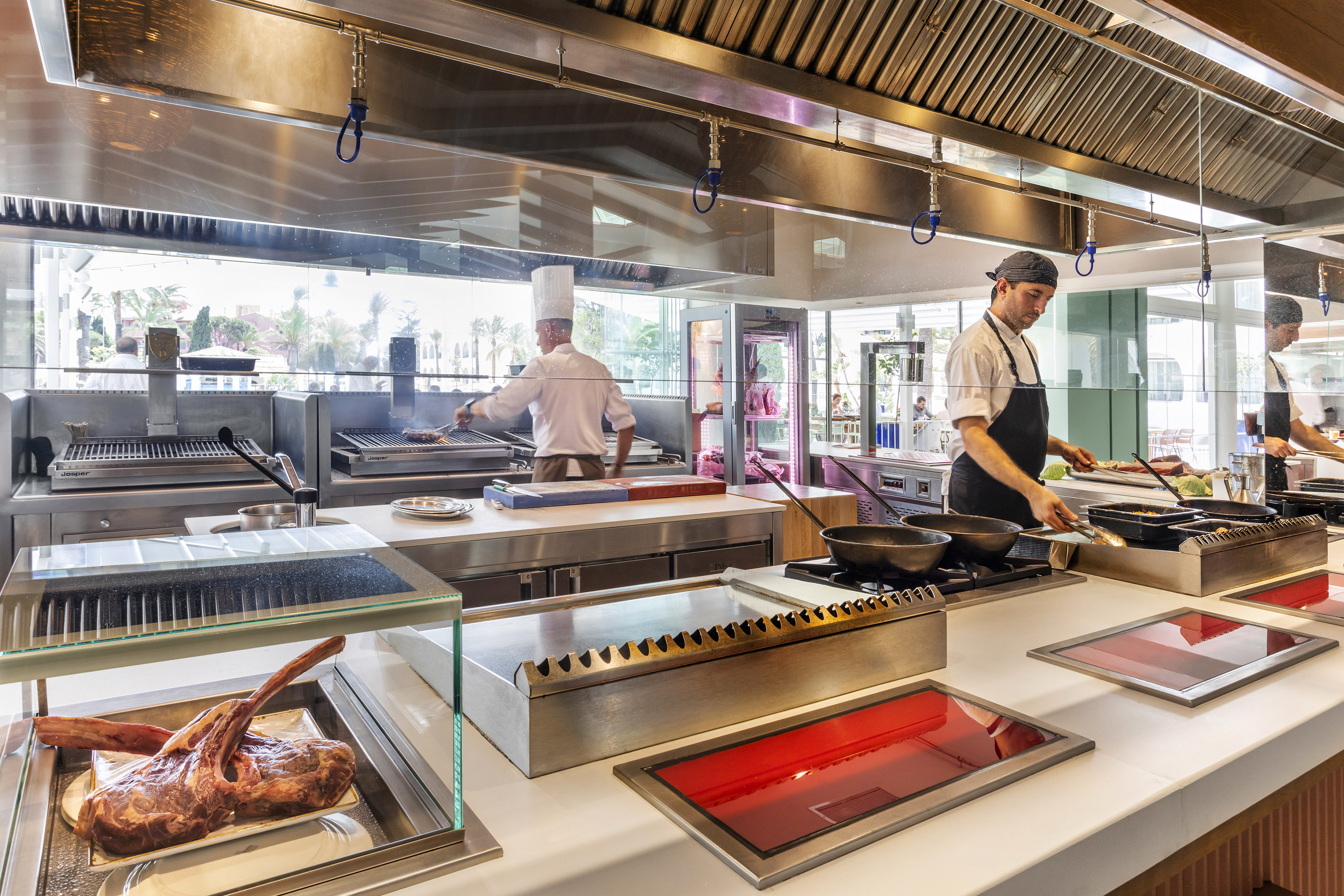 Modern open kitchen with chefs grilling, showcasing raw meat and cooking stations.