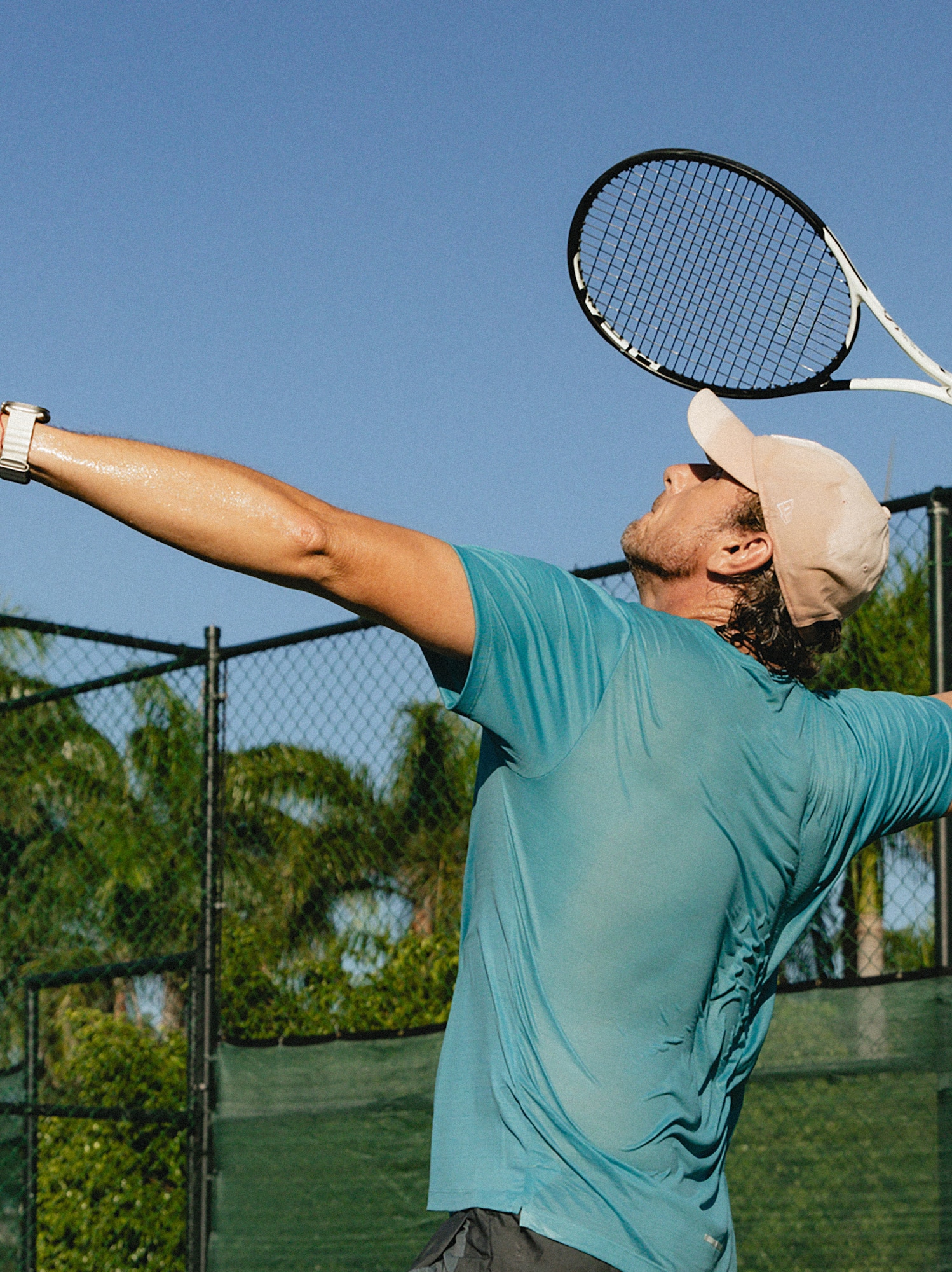 Male tennis player serving overhead on a sunny outdoor court.