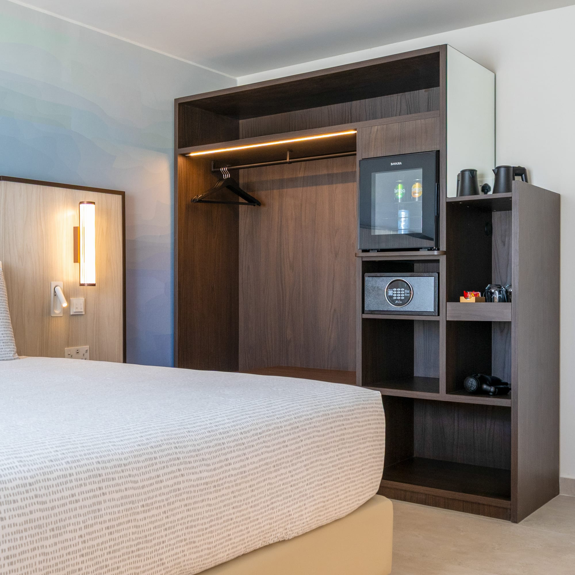 Modern hotel bedroom with built-in wardrobe, minibar, safe, and patterned bed.