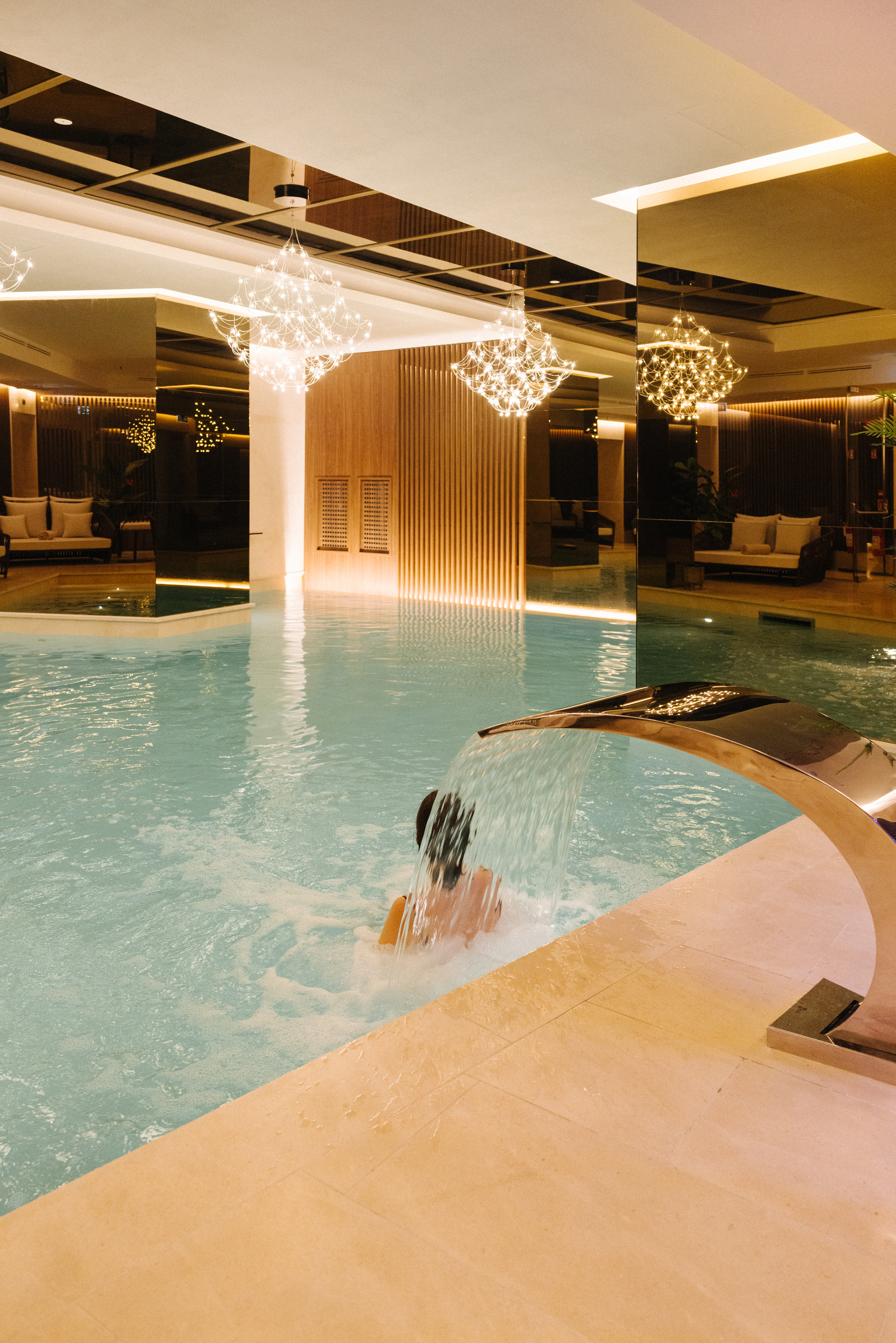 Luxury indoor spa pool with person under a waterfall, modern chandeliers.