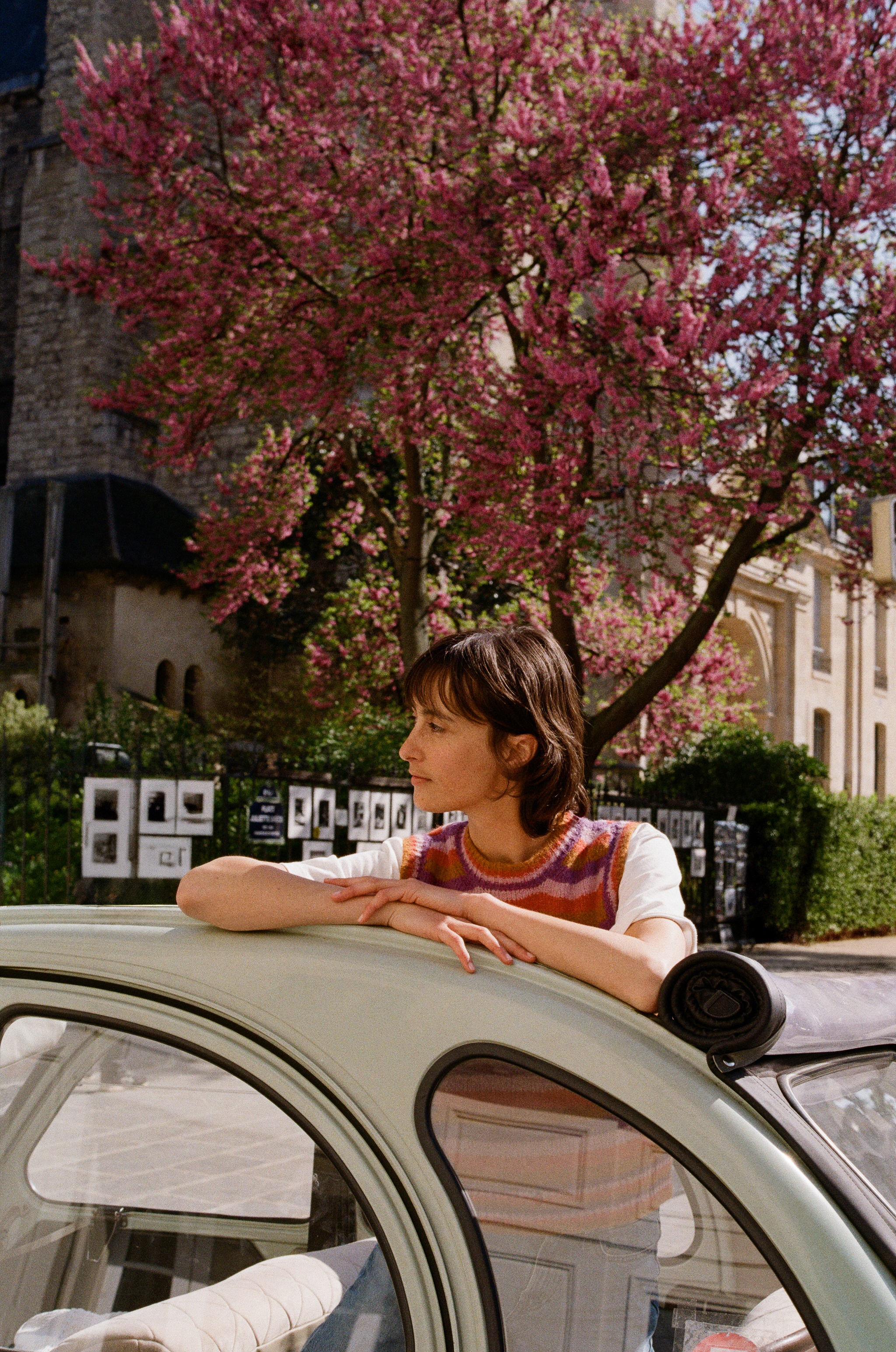 Mujer con chaleco, coche clásico verde, árbol rosa florecido en París.