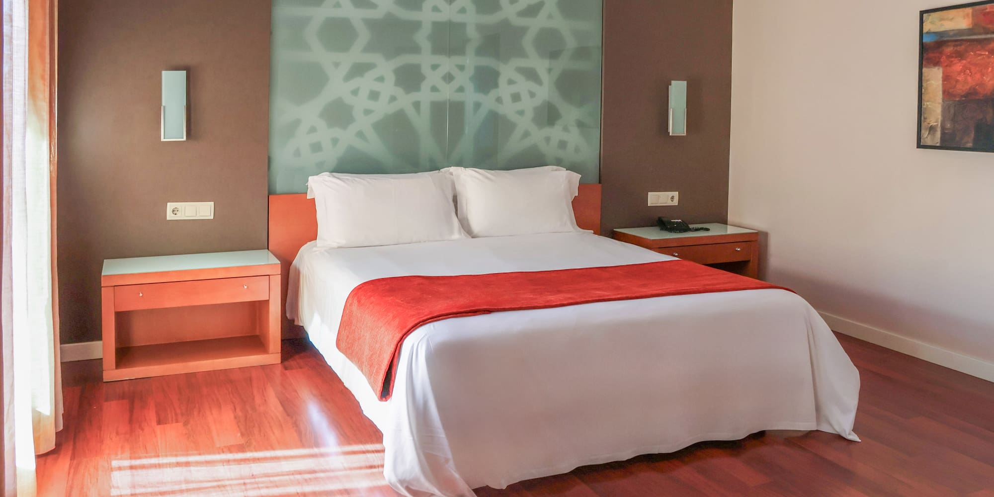 Modern hotel room with bed, red bed runner, patterned wall, and wood flooring.