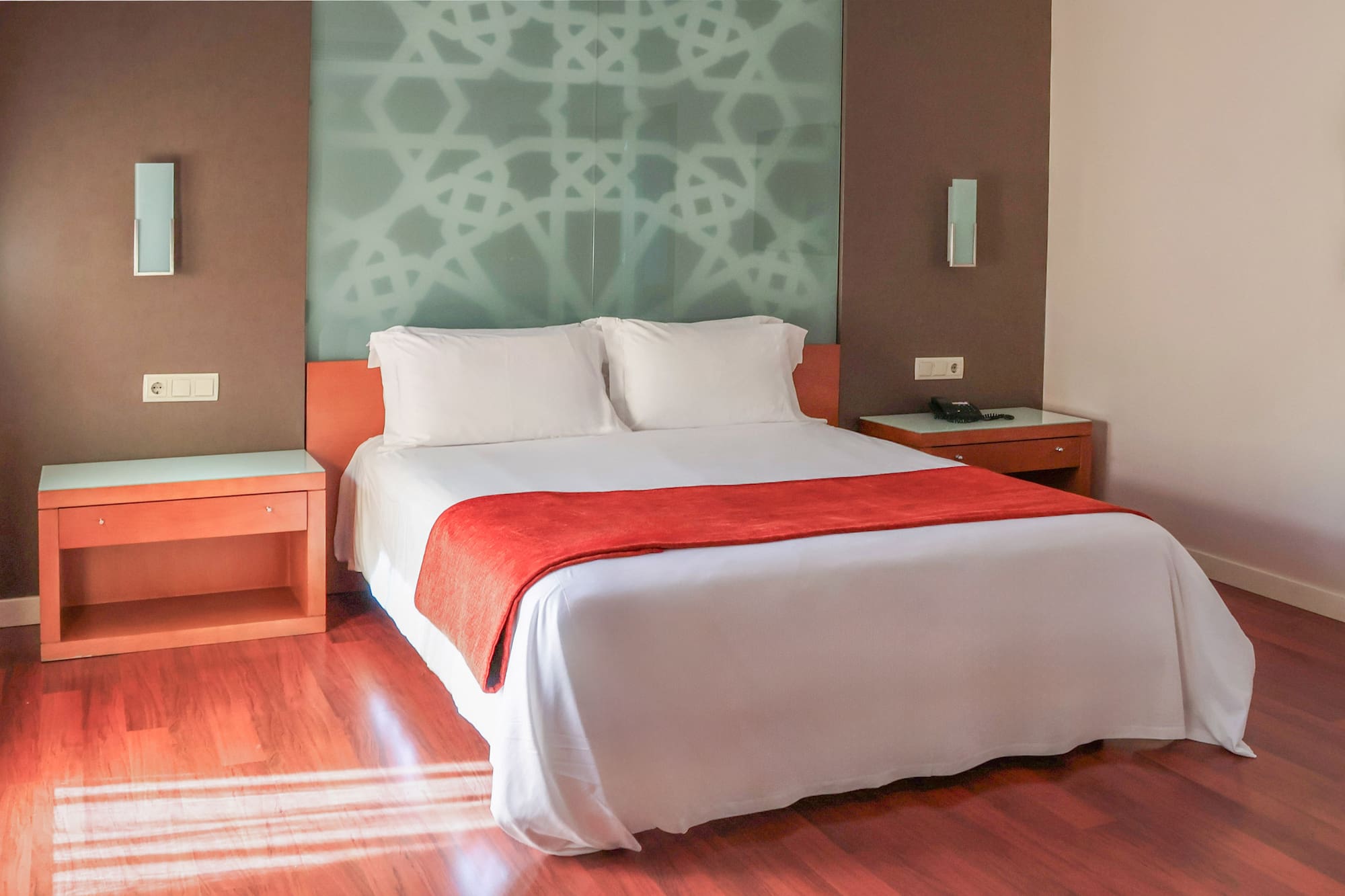 Modern hotel room with bed, red bed runner, patterned wall, and wood flooring.