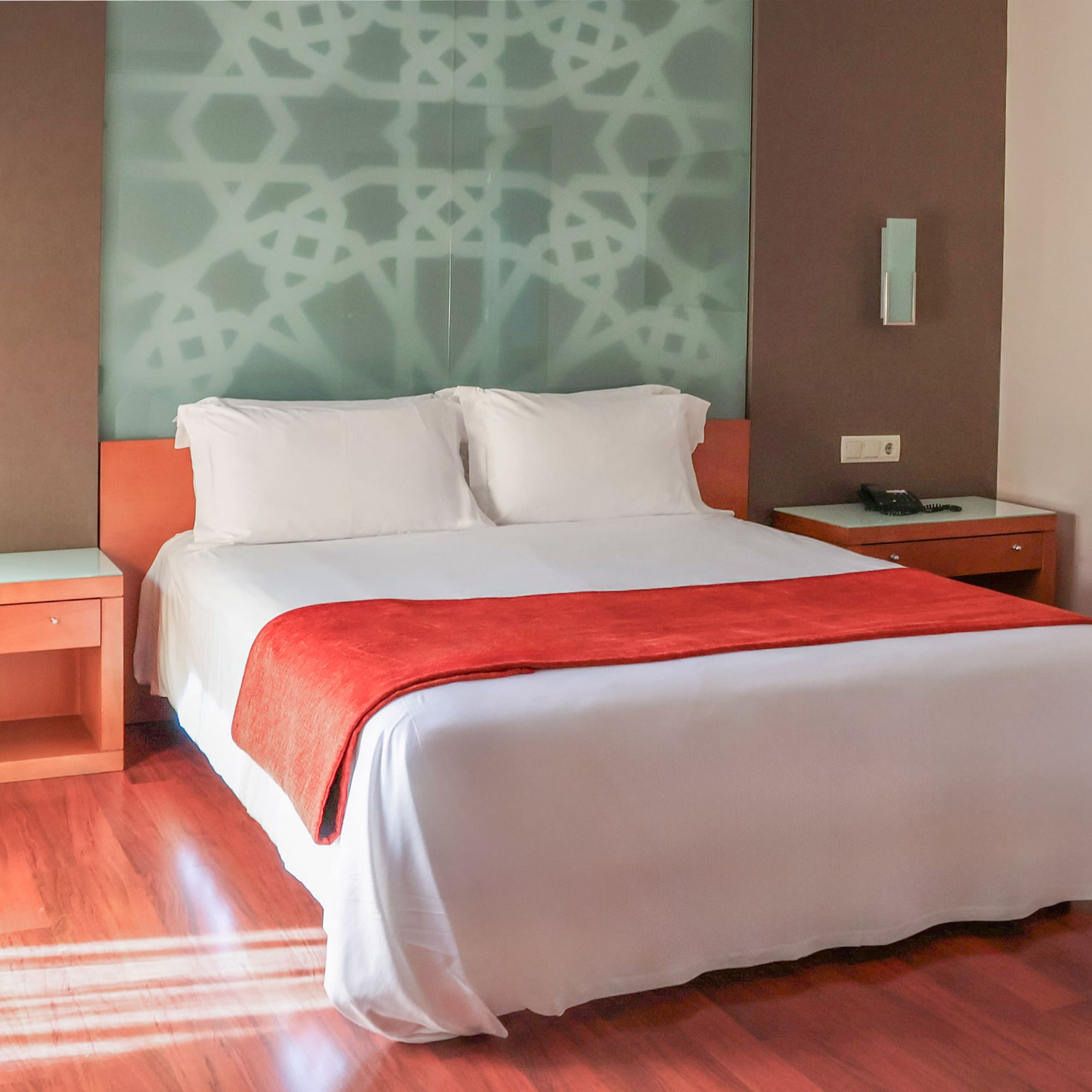 Modern hotel room with bed, red bed runner, patterned wall, and wood flooring.
