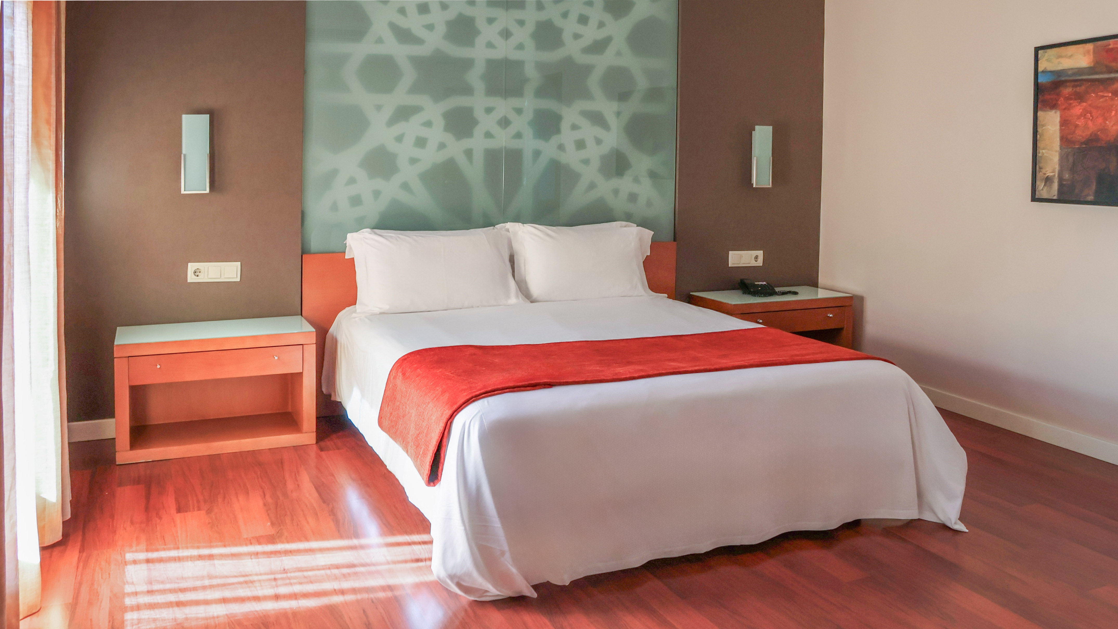 Modern hotel room with bed, red bed runner, patterned wall, and wood flooring.