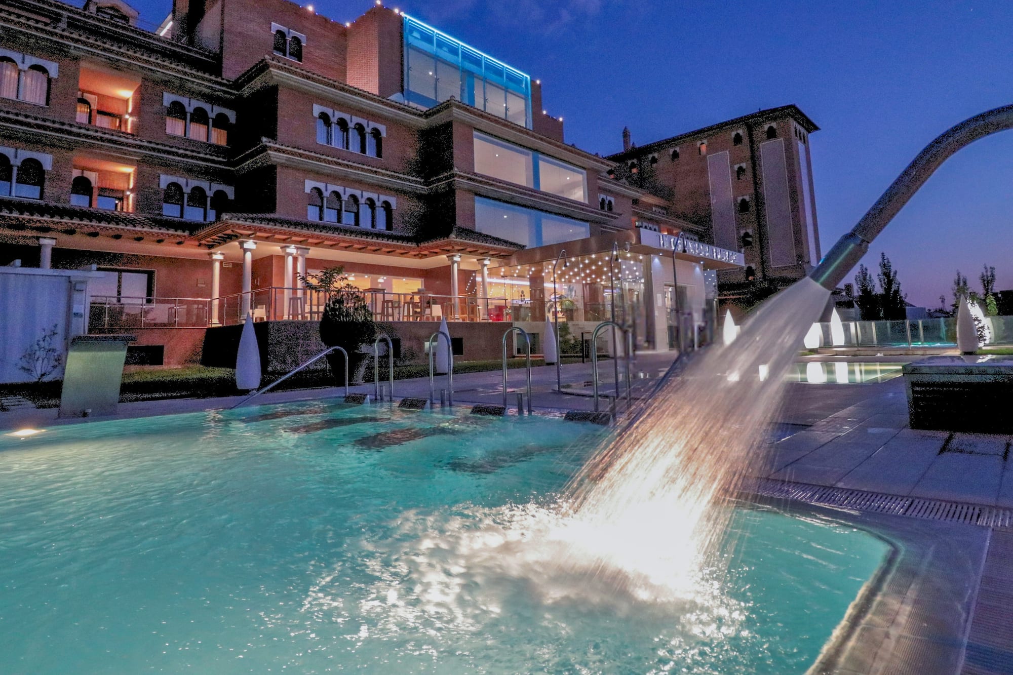 Luxury hotel outdoor pool at night with splashing water fountain.