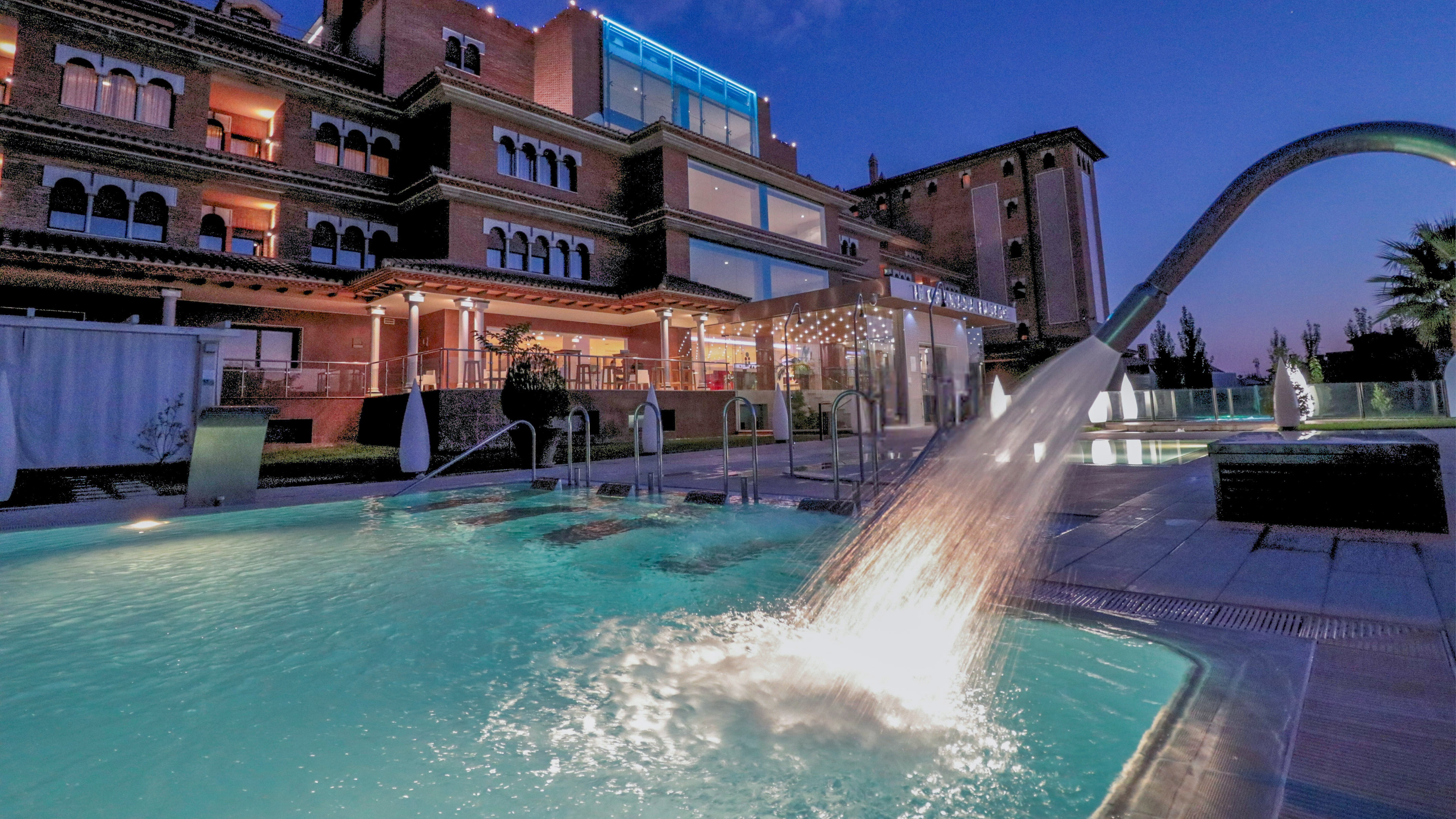 Luxury hotel outdoor pool at night with splashing water fountain.
