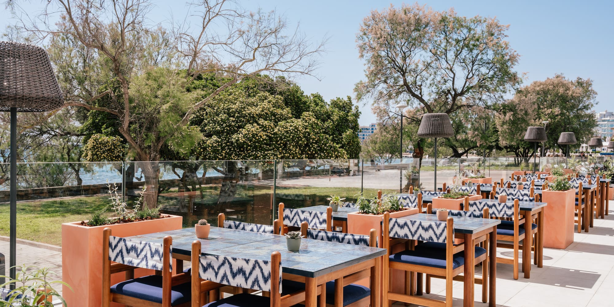 Sunny outdoor restaurant patio with blue tables, patterned chairs, sea view.