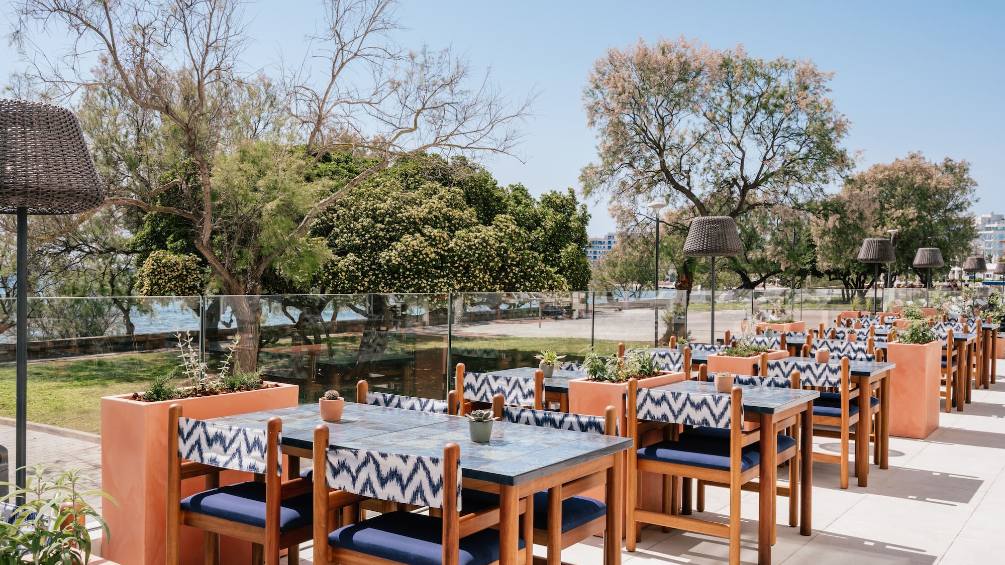 Sunny outdoor restaurant patio with blue tables, patterned chairs, sea view.